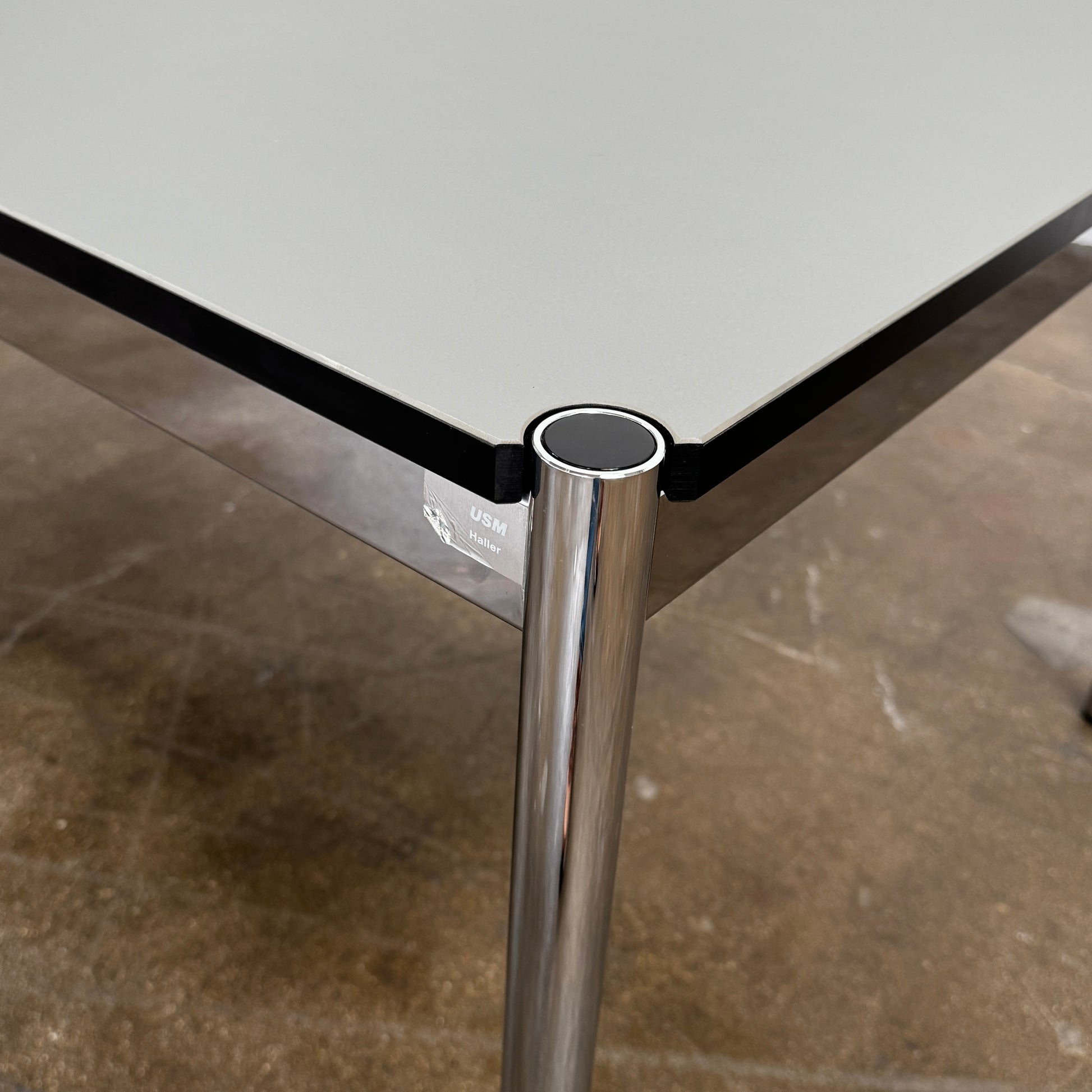 A modern mesh-back office chair sits at a USM Haller Work Table by USM Haller, which has a minimalist design and metal legs, placed on a concrete floor with a white background.