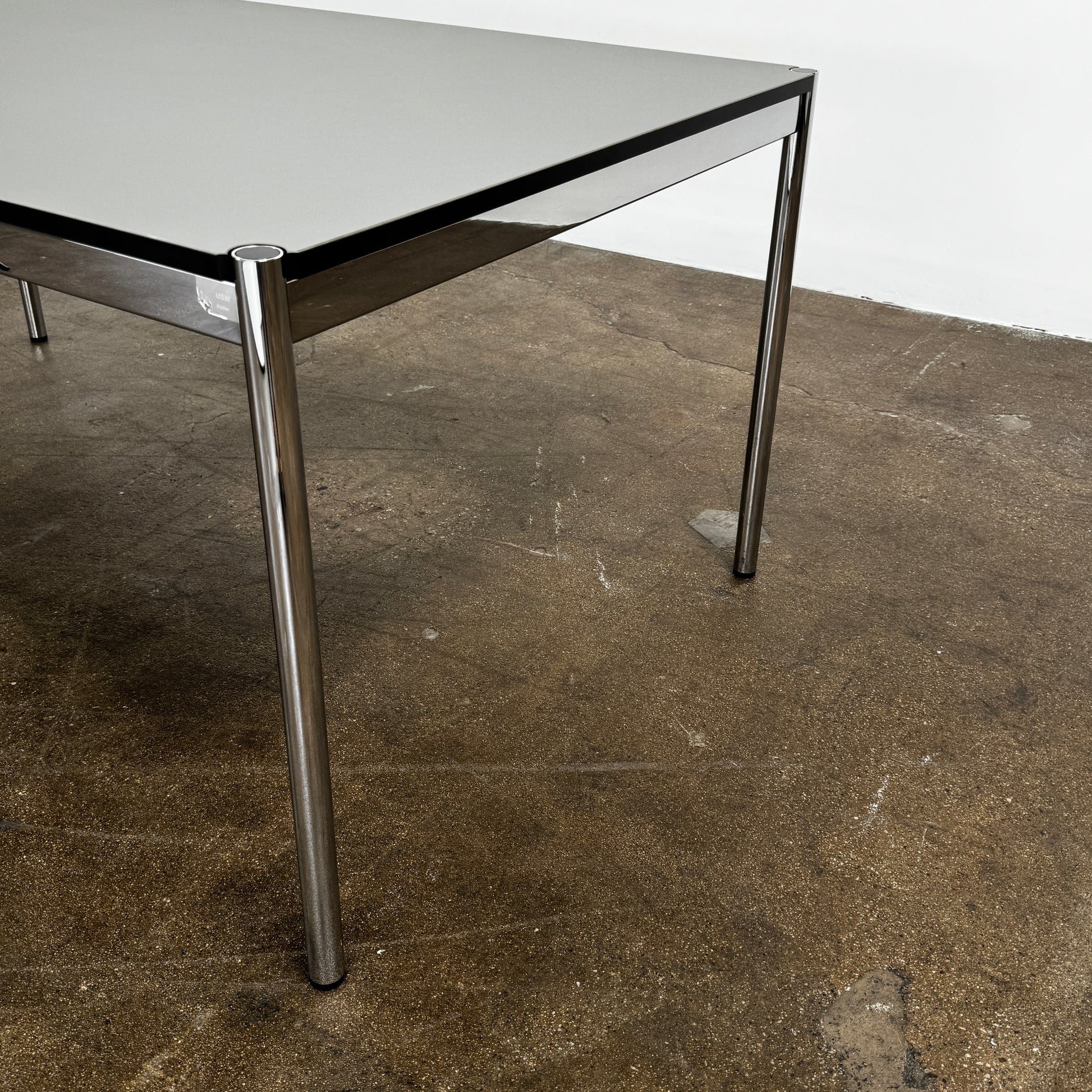 A modern mesh-back office chair sits at a USM Haller Work Table by USM Haller, which has a minimalist design and metal legs, placed on a concrete floor with a white background.