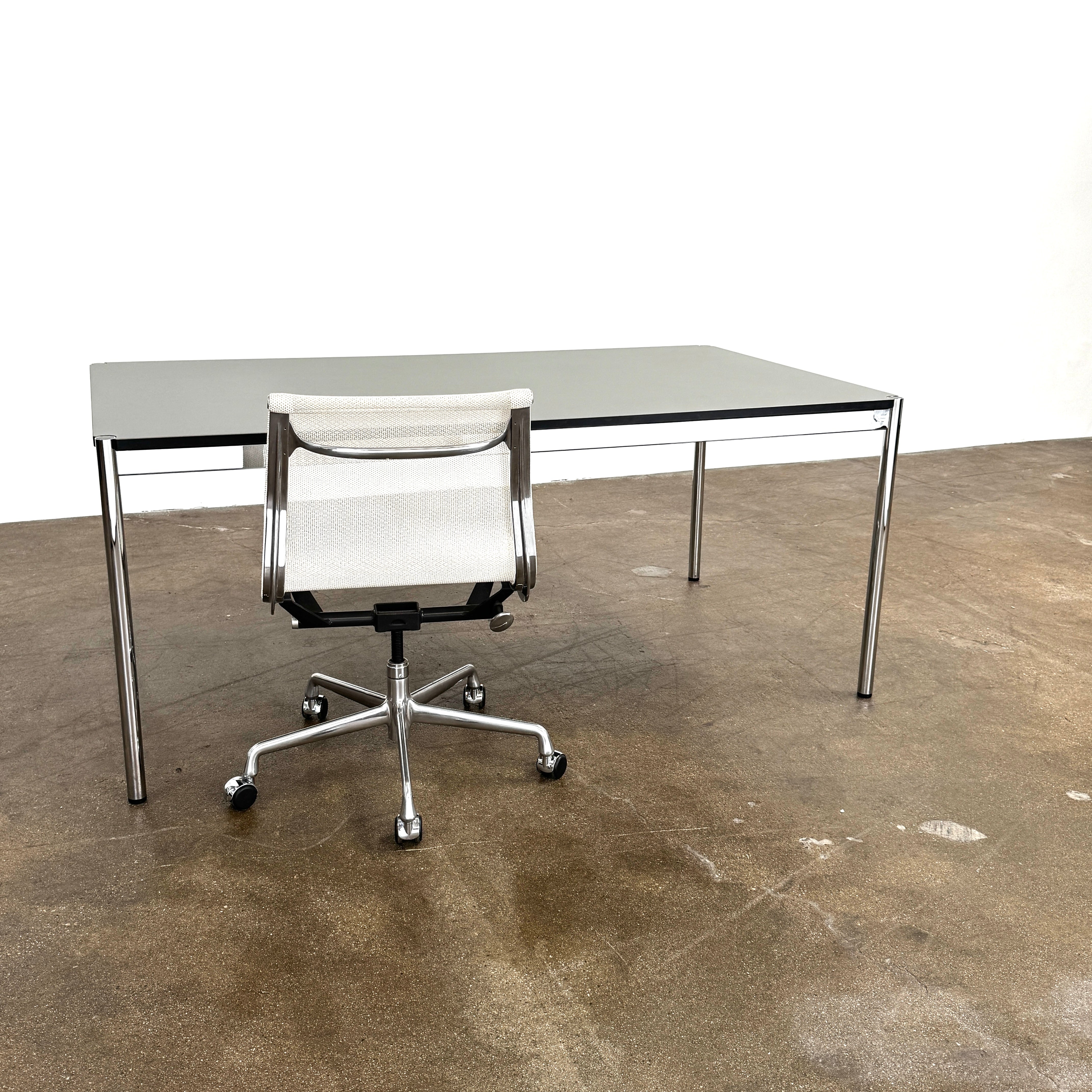A modern mesh-back office chair sits at a USM Haller Work Table by USM Haller, which has a minimalist design and metal legs, placed on a concrete floor with a white background.