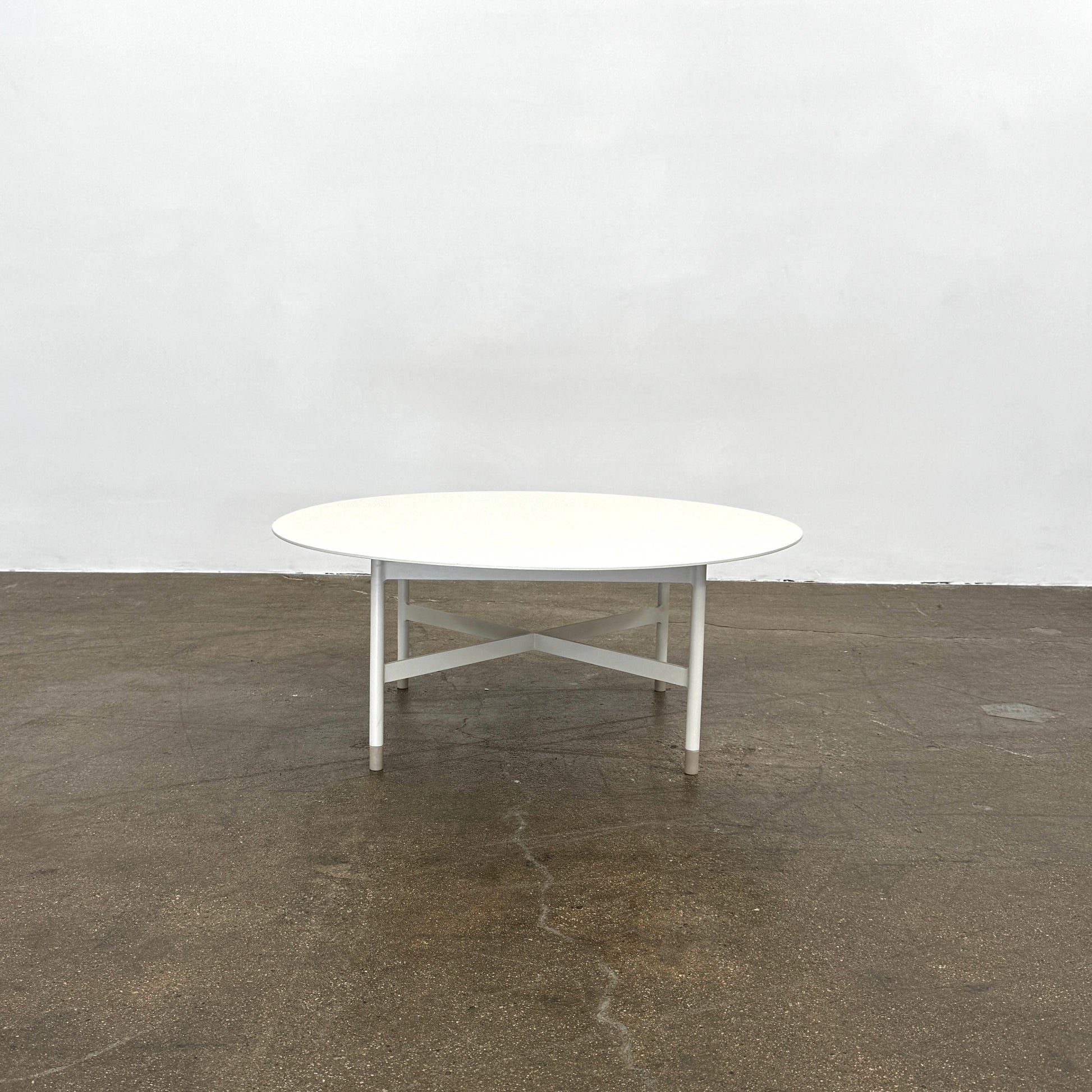 The MillerKnoll Sommer Outdoor Coffee Table for DWR is a round, white table with four legs and an X-shaped base, ideal for minimalist outdoor spaces. Brand: MillerKnoll. Set against a plain white background.