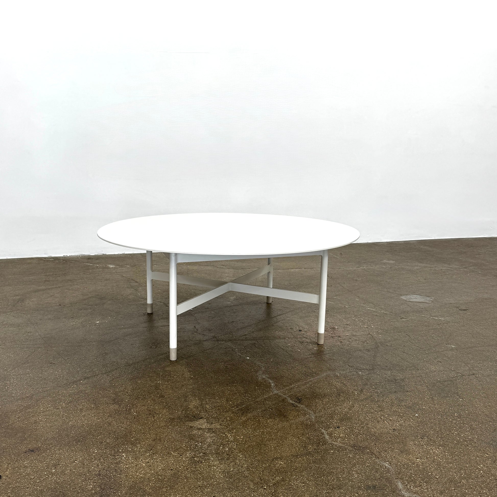 The MillerKnoll Sommer Outdoor Coffee Table for DWR is a round, white table with four legs and an X-shaped base, ideal for minimalist outdoor spaces. Brand: MillerKnoll. Set against a plain white background.