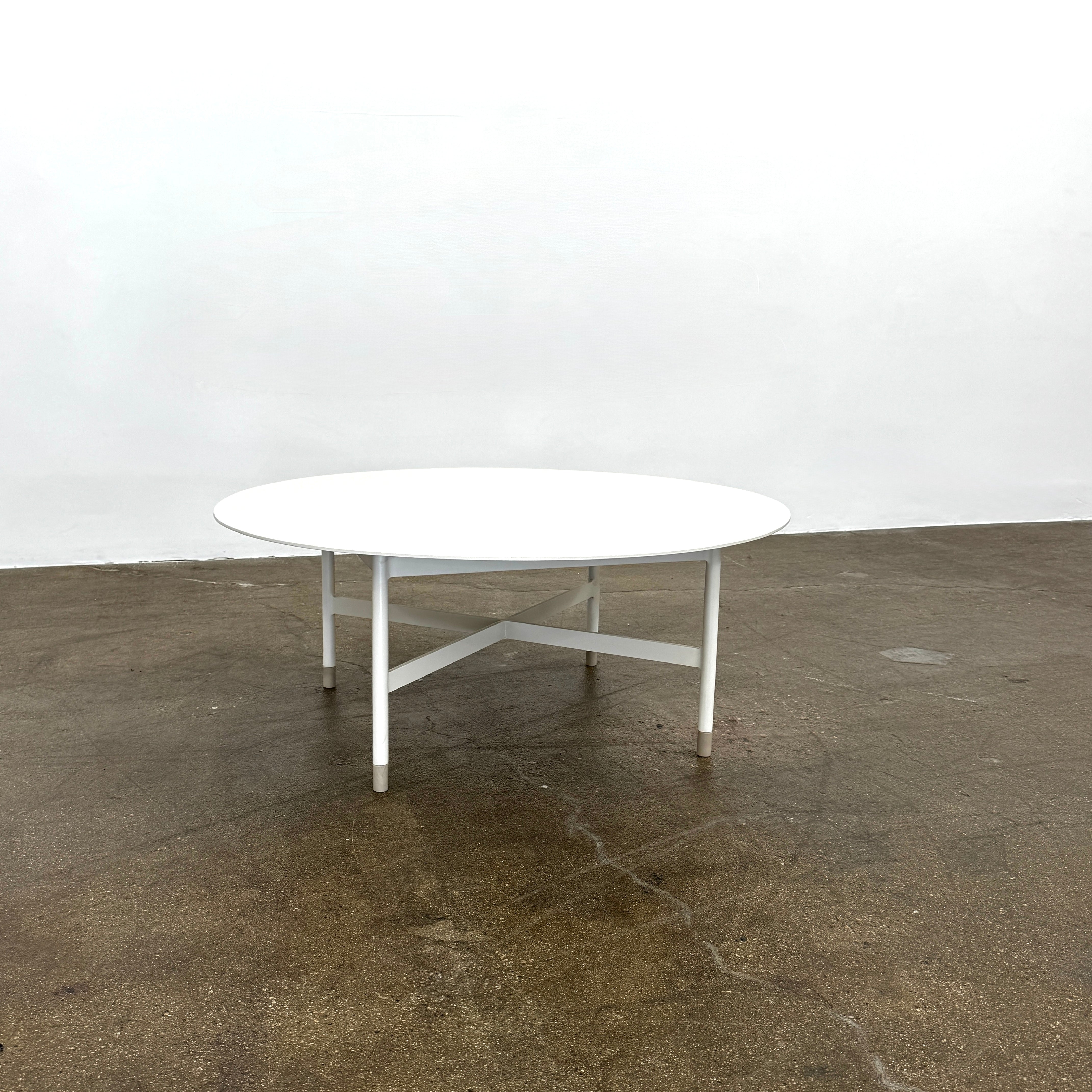 The MillerKnoll Sommer Outdoor Coffee Table for DWR is a round, white table with four legs and an X-shaped base, ideal for minimalist outdoor spaces. Brand: MillerKnoll. Set against a plain white background.