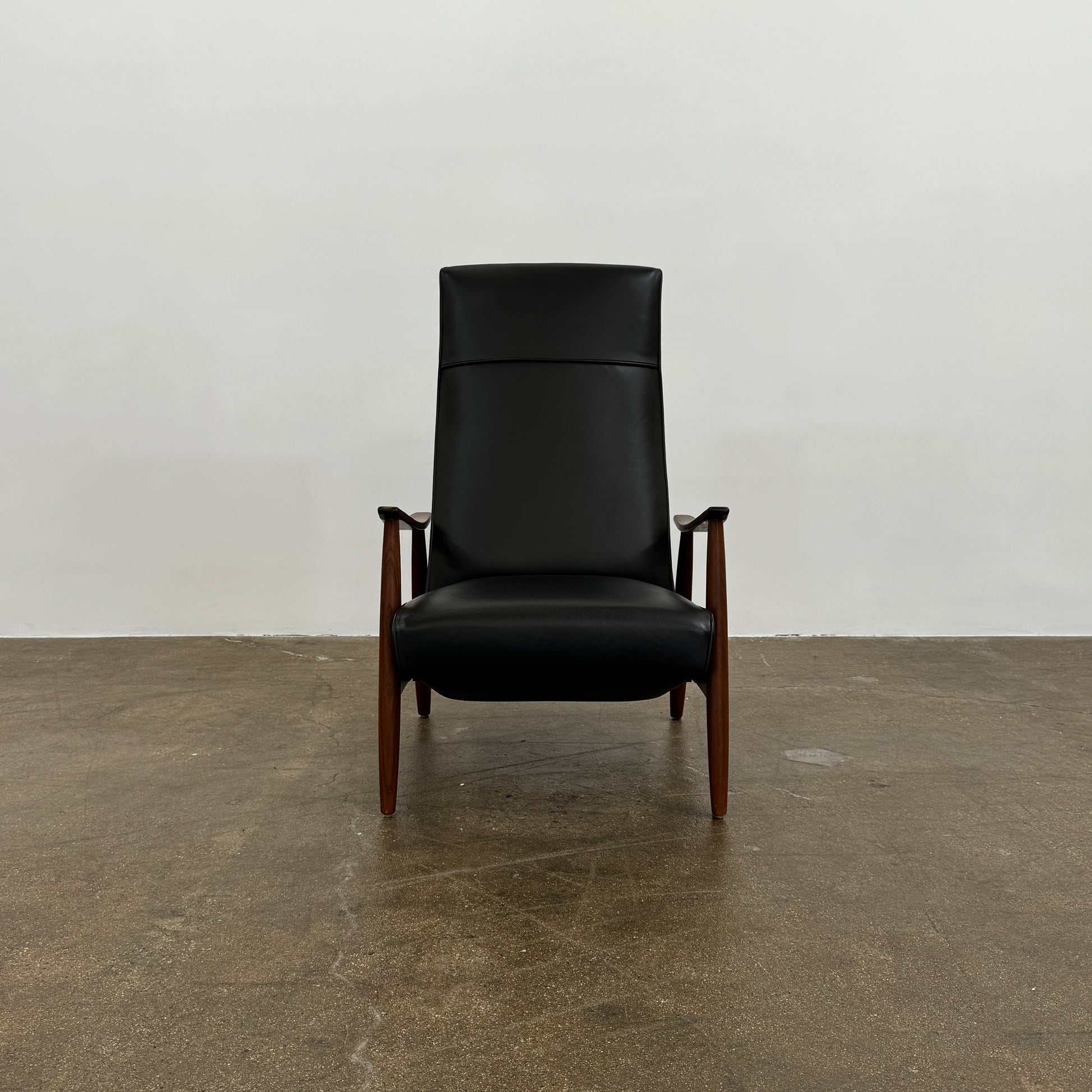 The Thayer Coggin Milo Baughman Recliner 74, featuring a black leather seat and solid walnut frame with wooden arms and legs, sits on a bare concrete floor against a plain white wall.