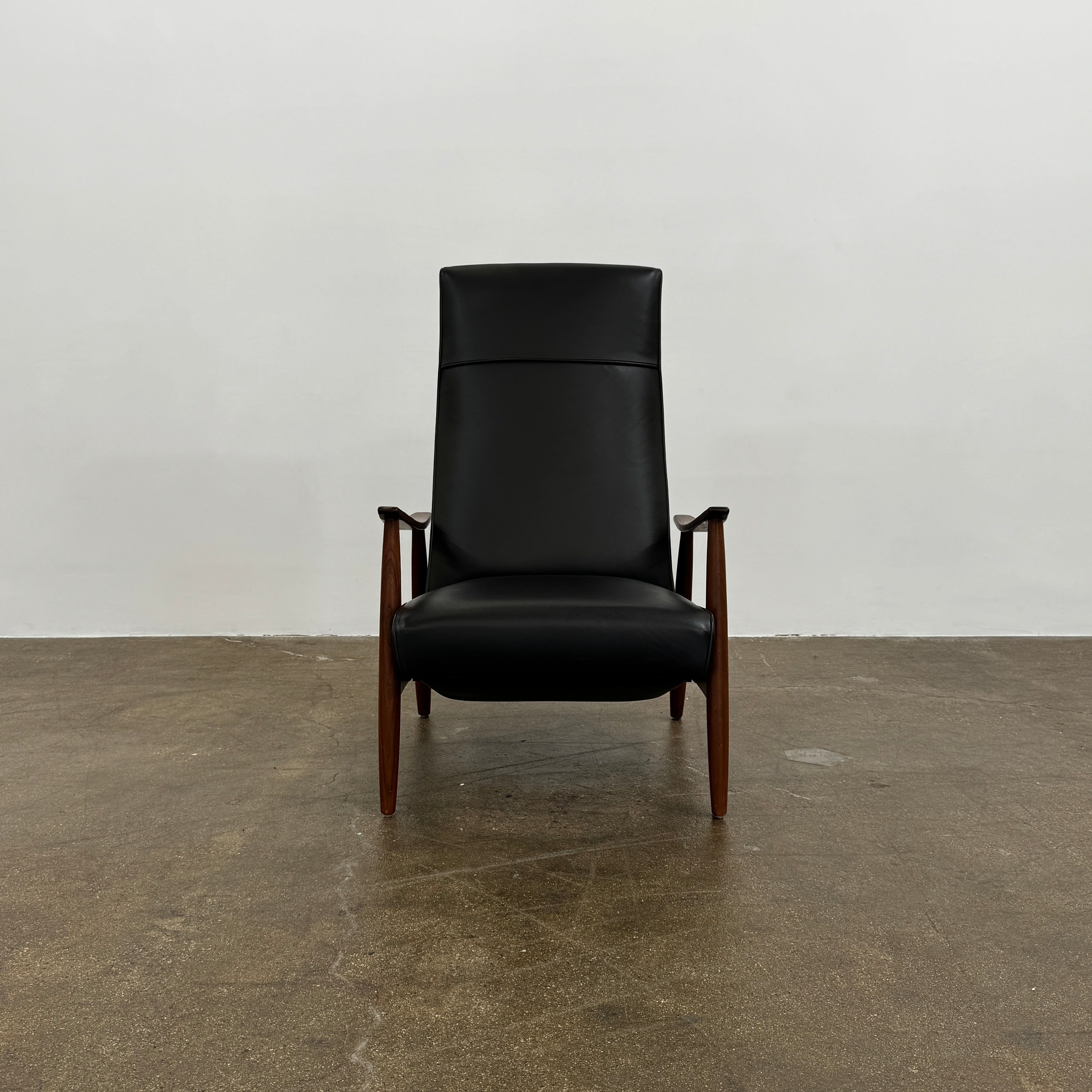 The Thayer Coggin Milo Baughman Recliner 74, featuring a black leather seat and solid walnut frame with wooden arms and legs, sits on a bare concrete floor against a plain white wall.