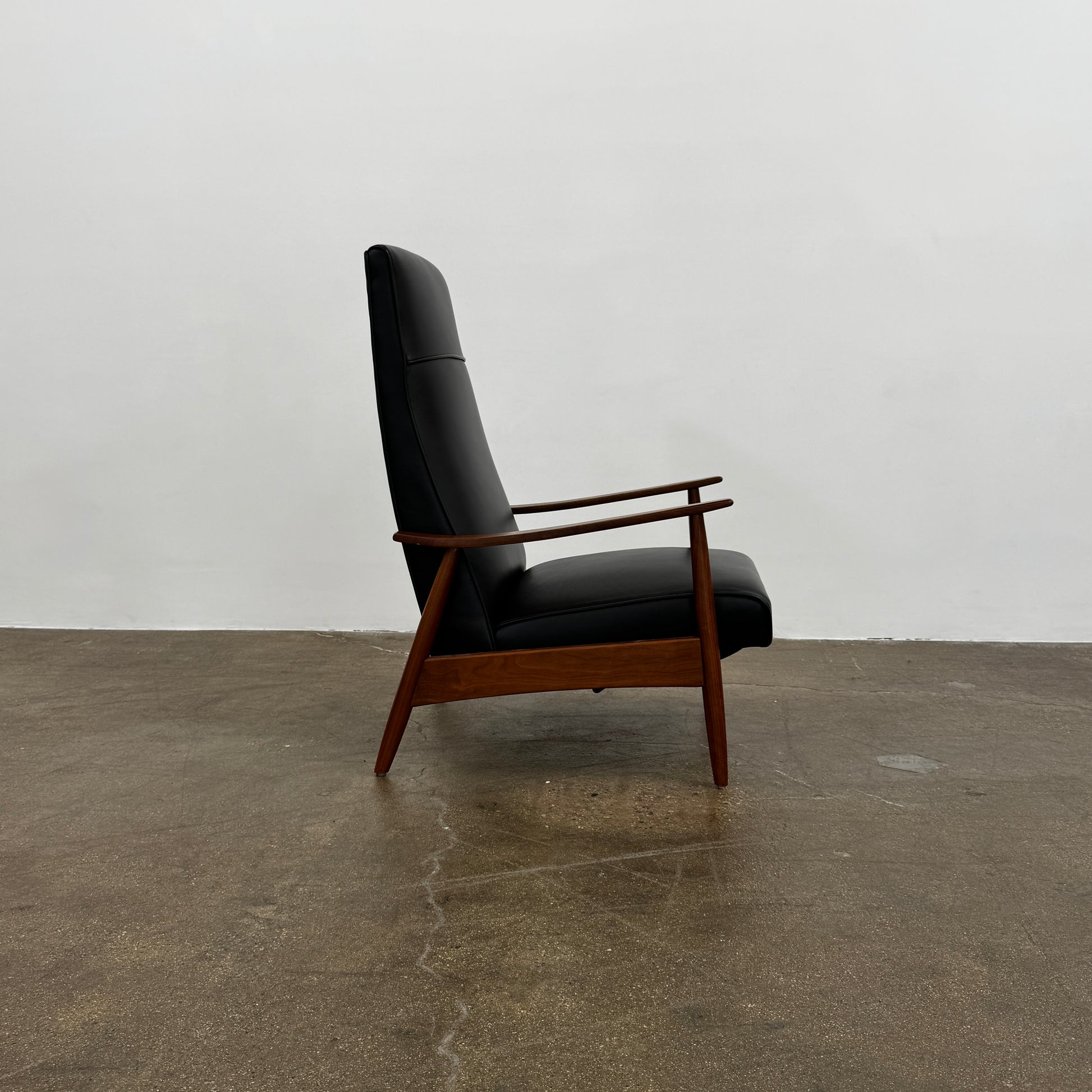 The Thayer Coggin Milo Baughman Recliner 74, featuring a black leather seat and solid walnut frame with wooden arms and legs, sits on a bare concrete floor against a plain white wall.