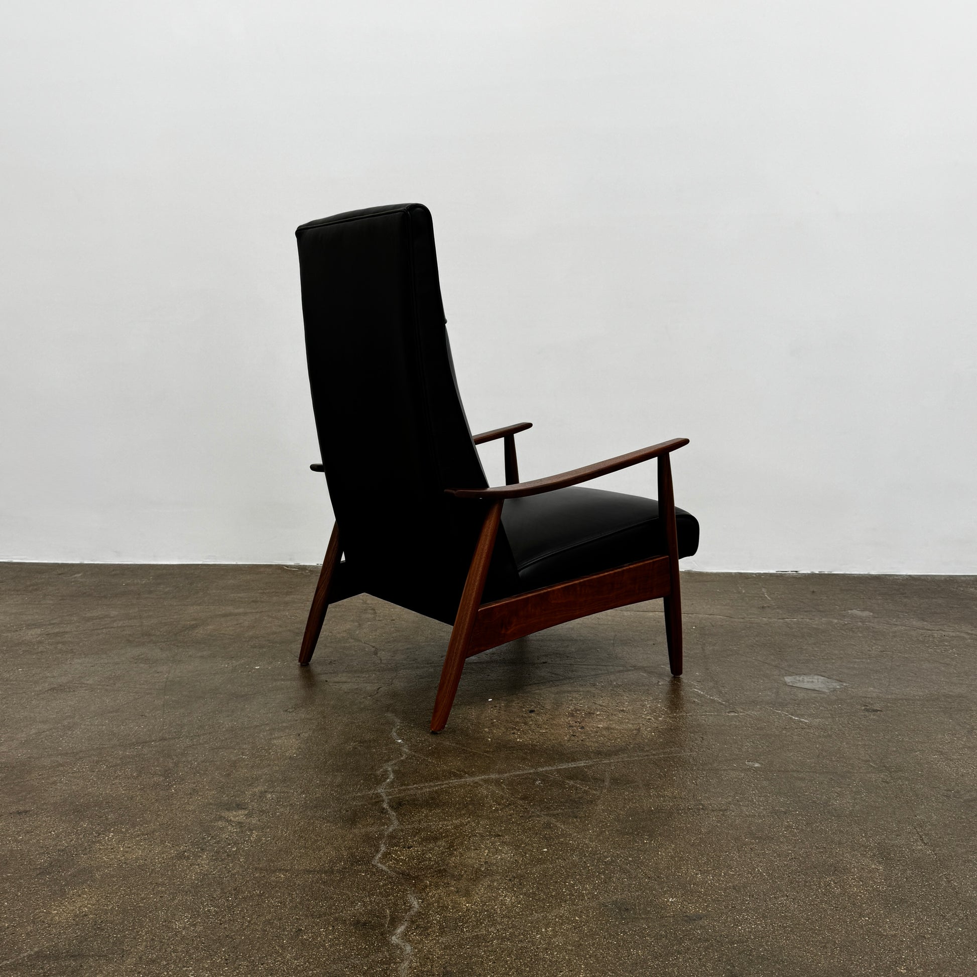 The Thayer Coggin Milo Baughman Recliner 74, featuring a black leather seat and solid walnut frame with wooden arms and legs, sits on a bare concrete floor against a plain white wall.