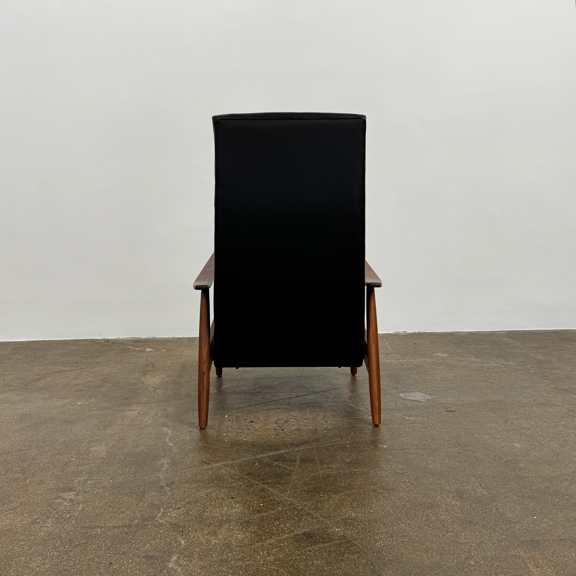 The Thayer Coggin Milo Baughman Recliner 74, featuring a black leather seat and solid walnut frame with wooden arms and legs, sits on a bare concrete floor against a plain white wall.