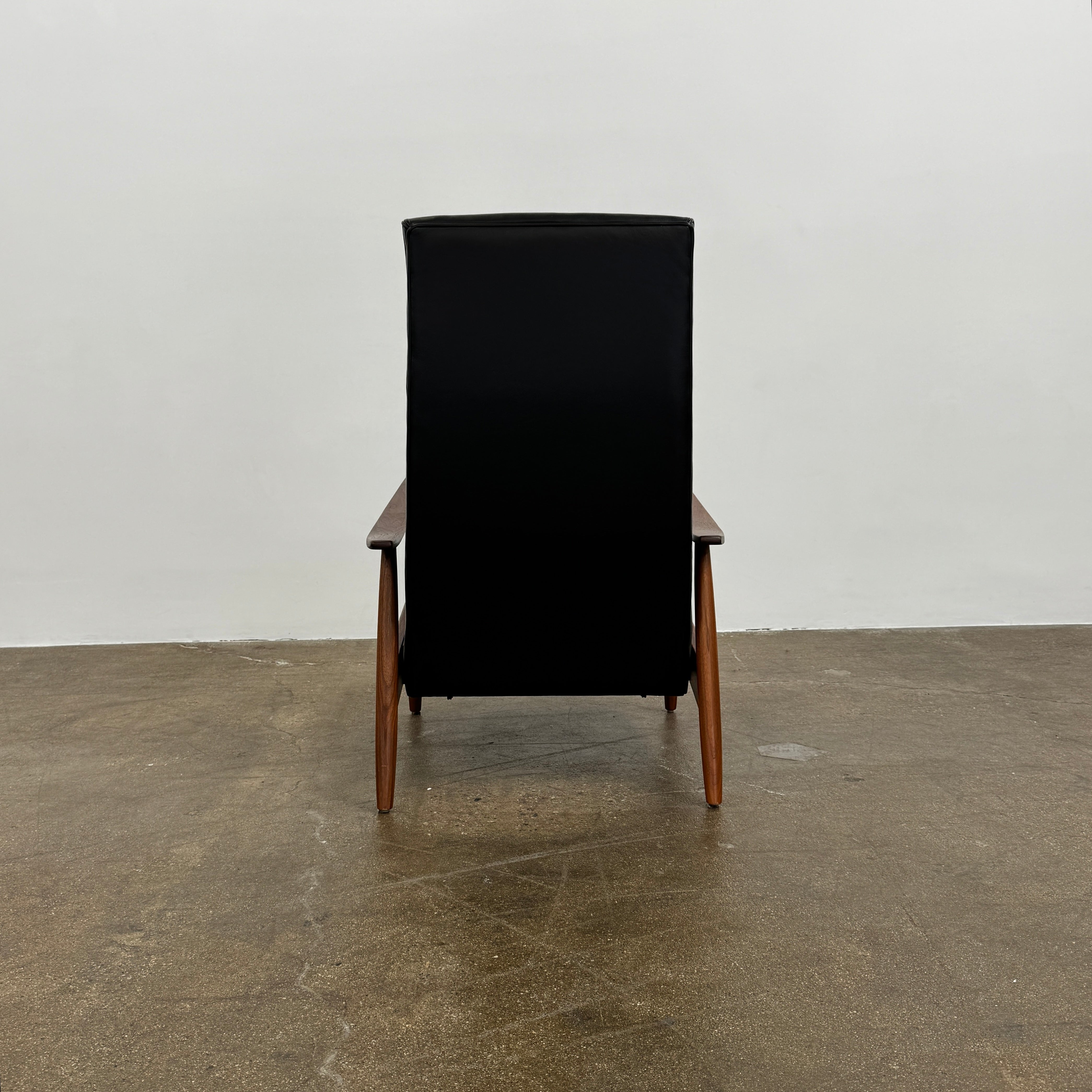 The Thayer Coggin Milo Baughman Recliner 74, featuring a black leather seat and solid walnut frame with wooden arms and legs, sits on a bare concrete floor against a plain white wall.