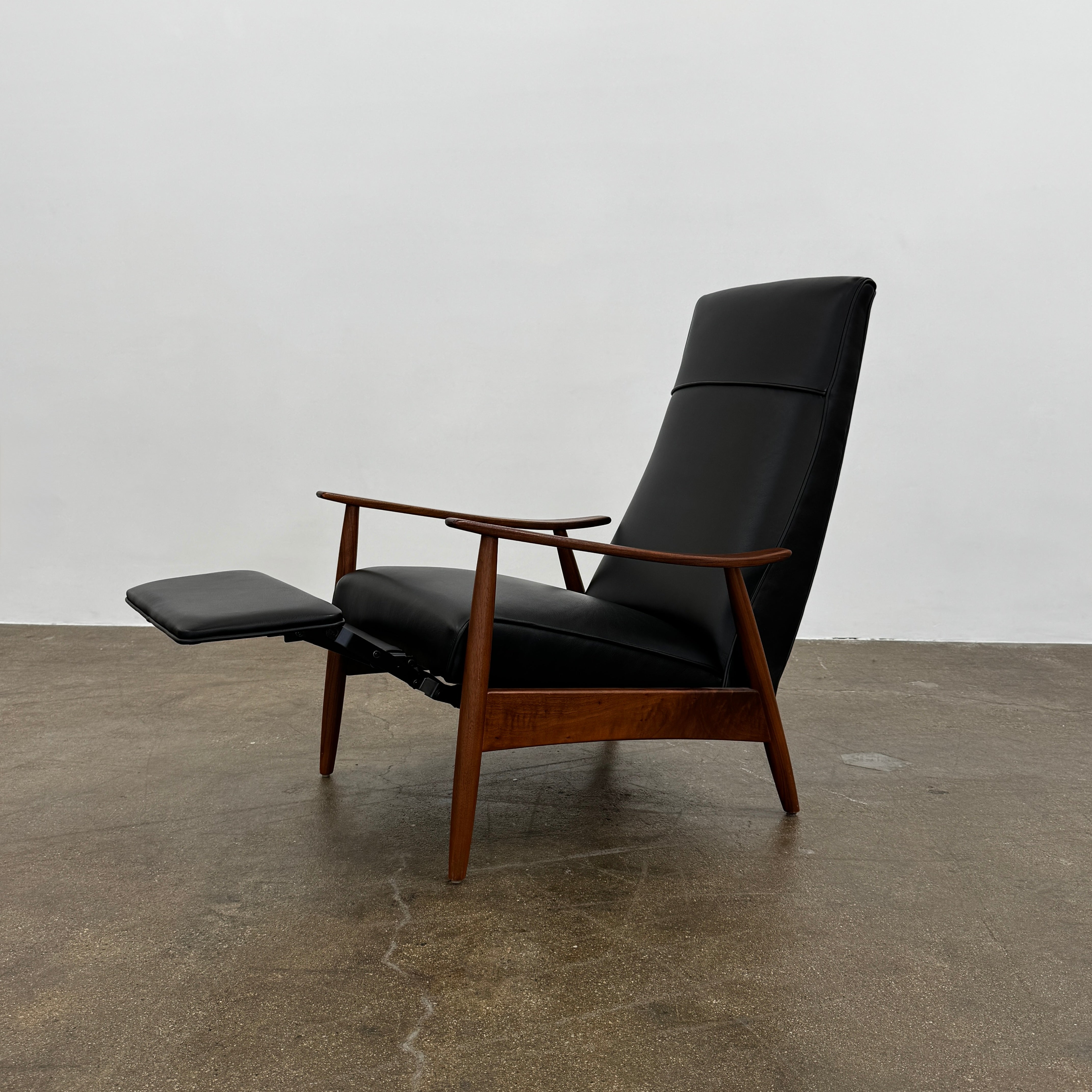 The Thayer Coggin Milo Baughman Recliner 74, featuring a black leather seat and solid walnut frame with wooden arms and legs, sits on a bare concrete floor against a plain white wall.