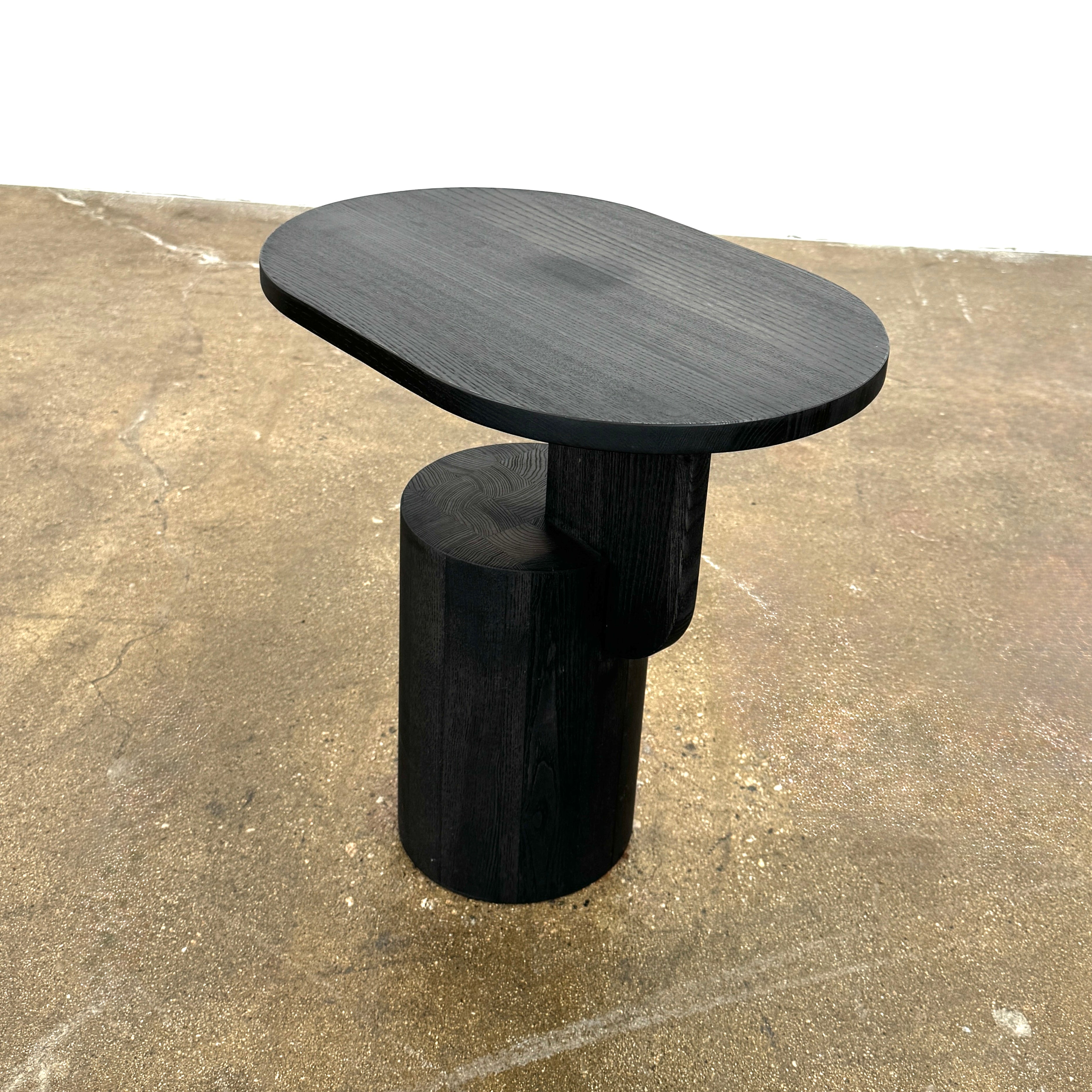 The Ferm Living Insert Side Table by Ferm Living features an oval dark wood top with a pedestal base, creating a striking accent on concrete floors and showcasing subtle Scandinavian design.