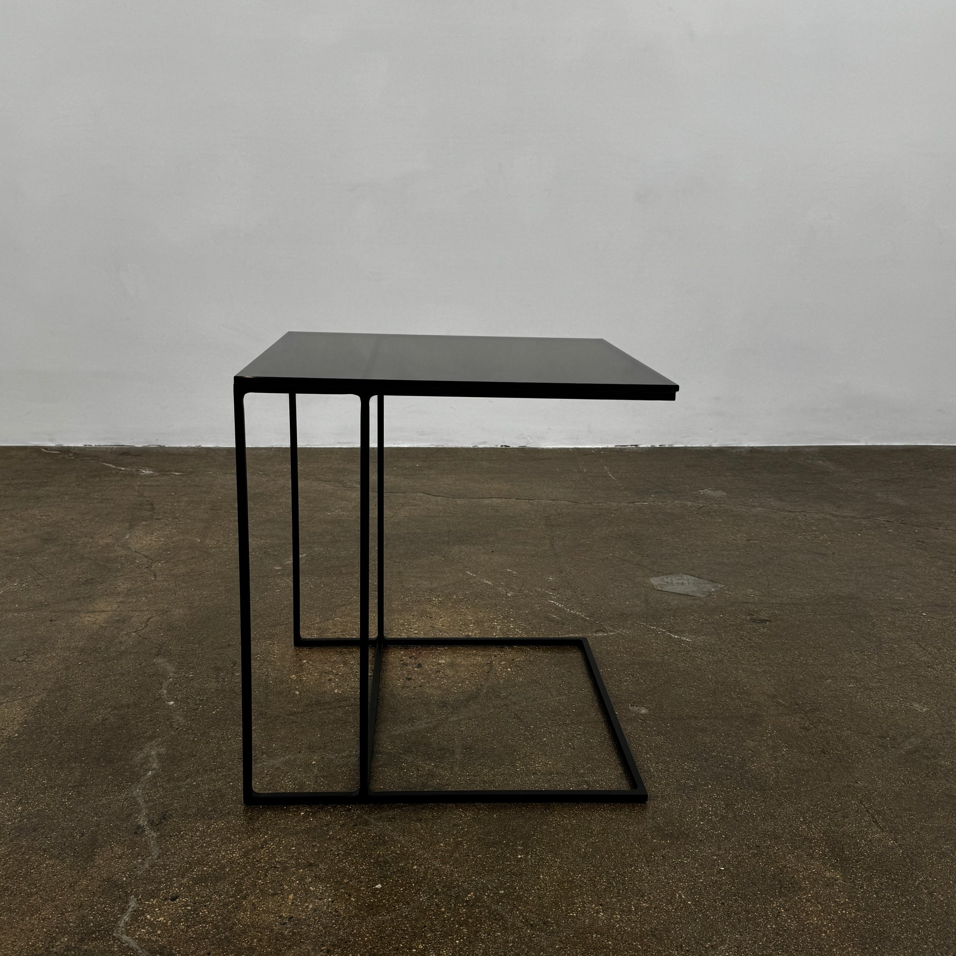 The Minotti Leger accent table by Minotti features a minimalist black metal frame with geometric lines and a flat rectangular bronze glass top, inspired by the cantilever style, shown against a plain background.