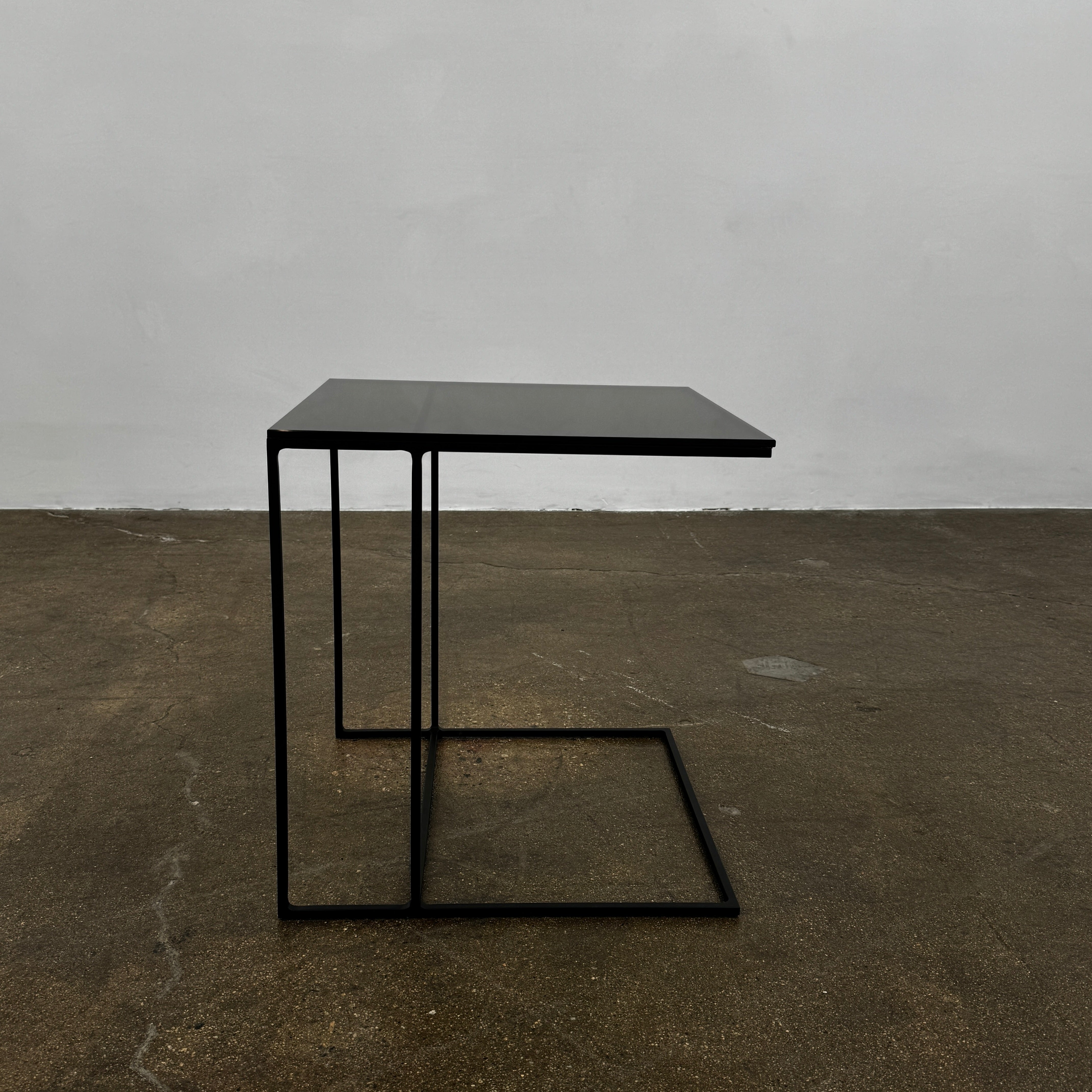 The Minotti Leger accent table by Minotti features a minimalist black metal frame with geometric lines and a flat rectangular bronze glass top, inspired by the cantilever style, shown against a plain background.