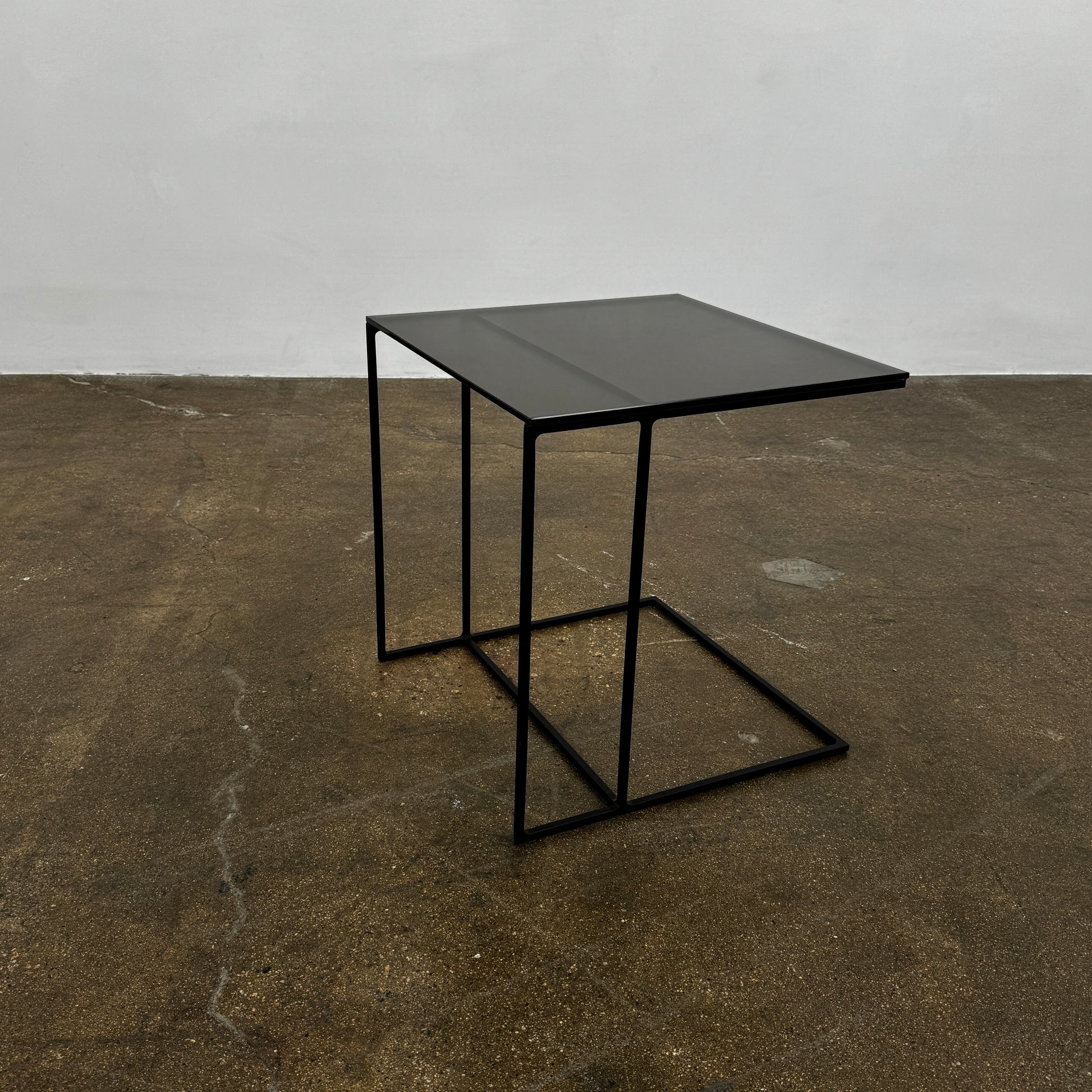 The Minotti Leger accent table by Minotti features a minimalist black metal frame with geometric lines and a flat rectangular bronze glass top, inspired by the cantilever style, shown against a plain background.
