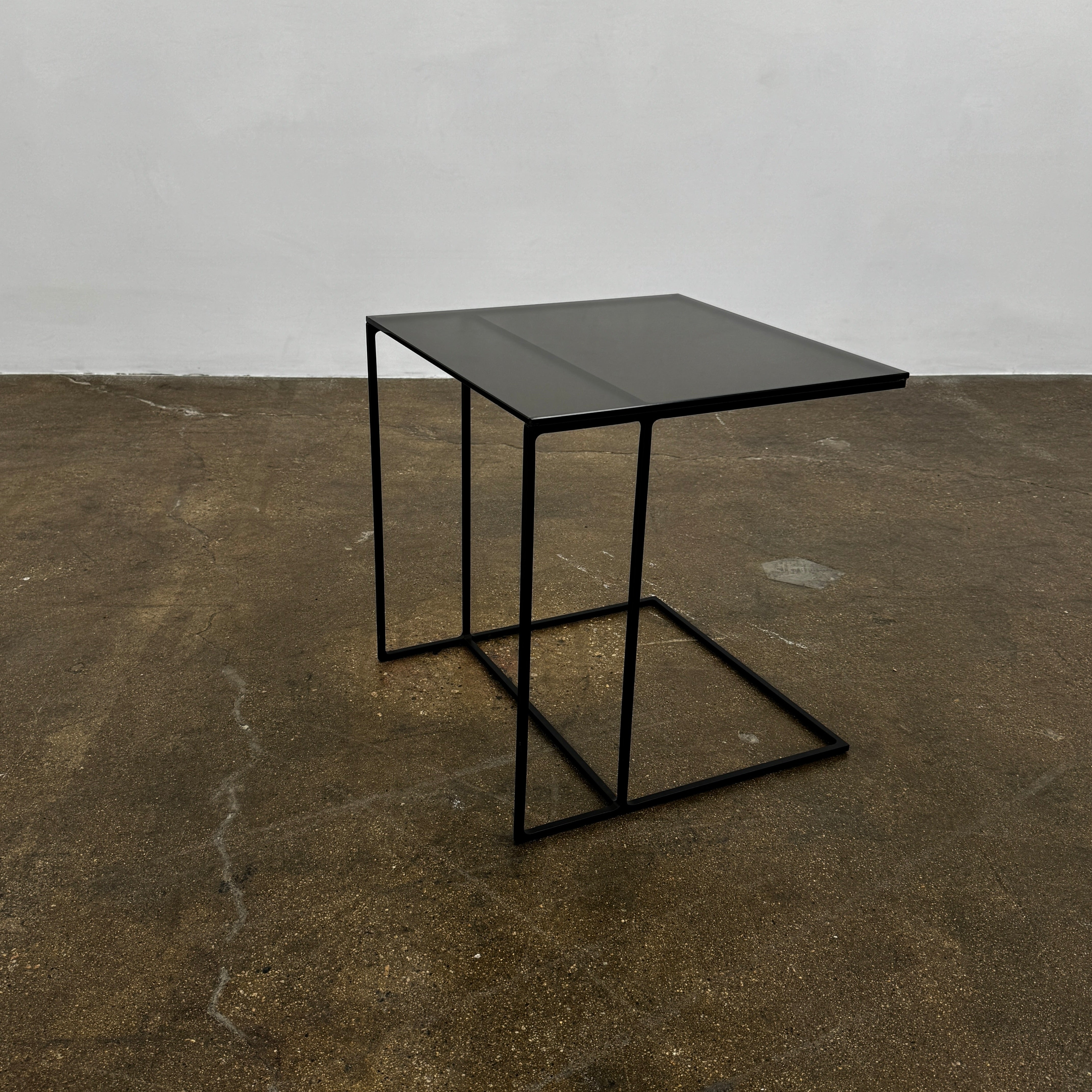 The Minotti Leger accent table by Minotti features a minimalist black metal frame with geometric lines and a flat rectangular bronze glass top, inspired by the cantilever style, shown against a plain background.
