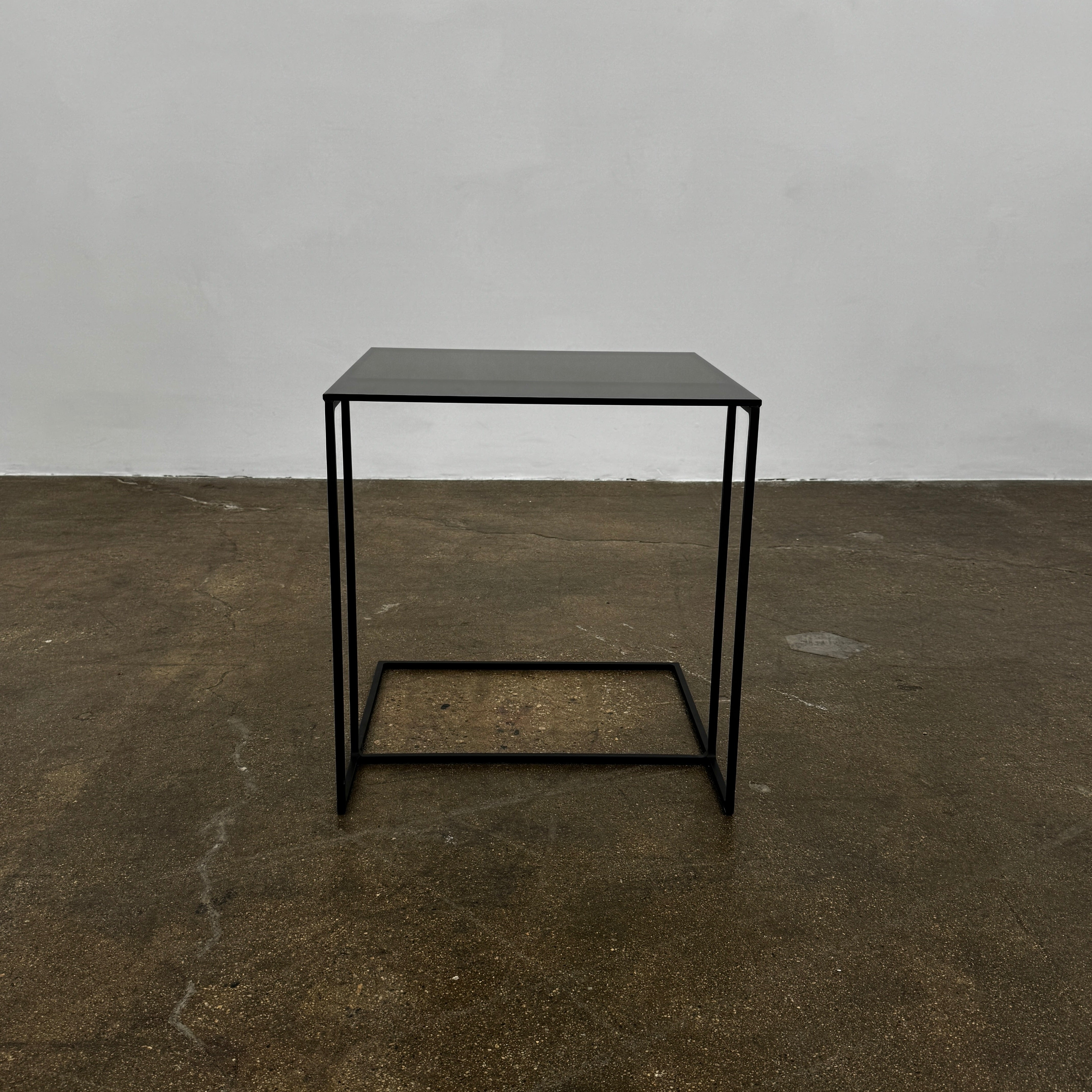 The Minotti Leger accent table by Minotti features a minimalist black metal frame with geometric lines and a flat rectangular bronze glass top, inspired by the cantilever style, shown against a plain background.
