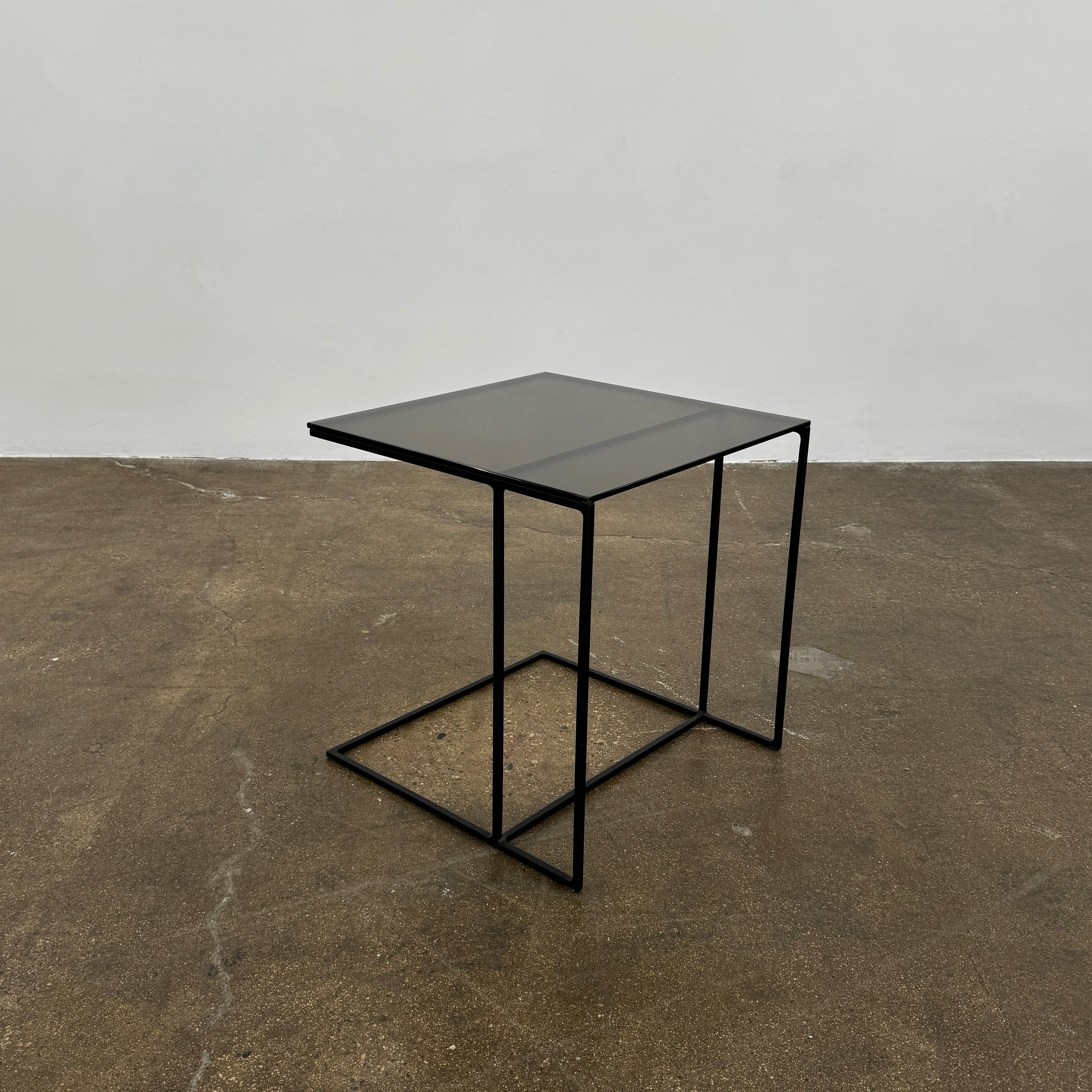 The Minotti Leger accent table by Minotti features a minimalist black metal frame with geometric lines and a flat rectangular bronze glass top, inspired by the cantilever style, shown against a plain background.