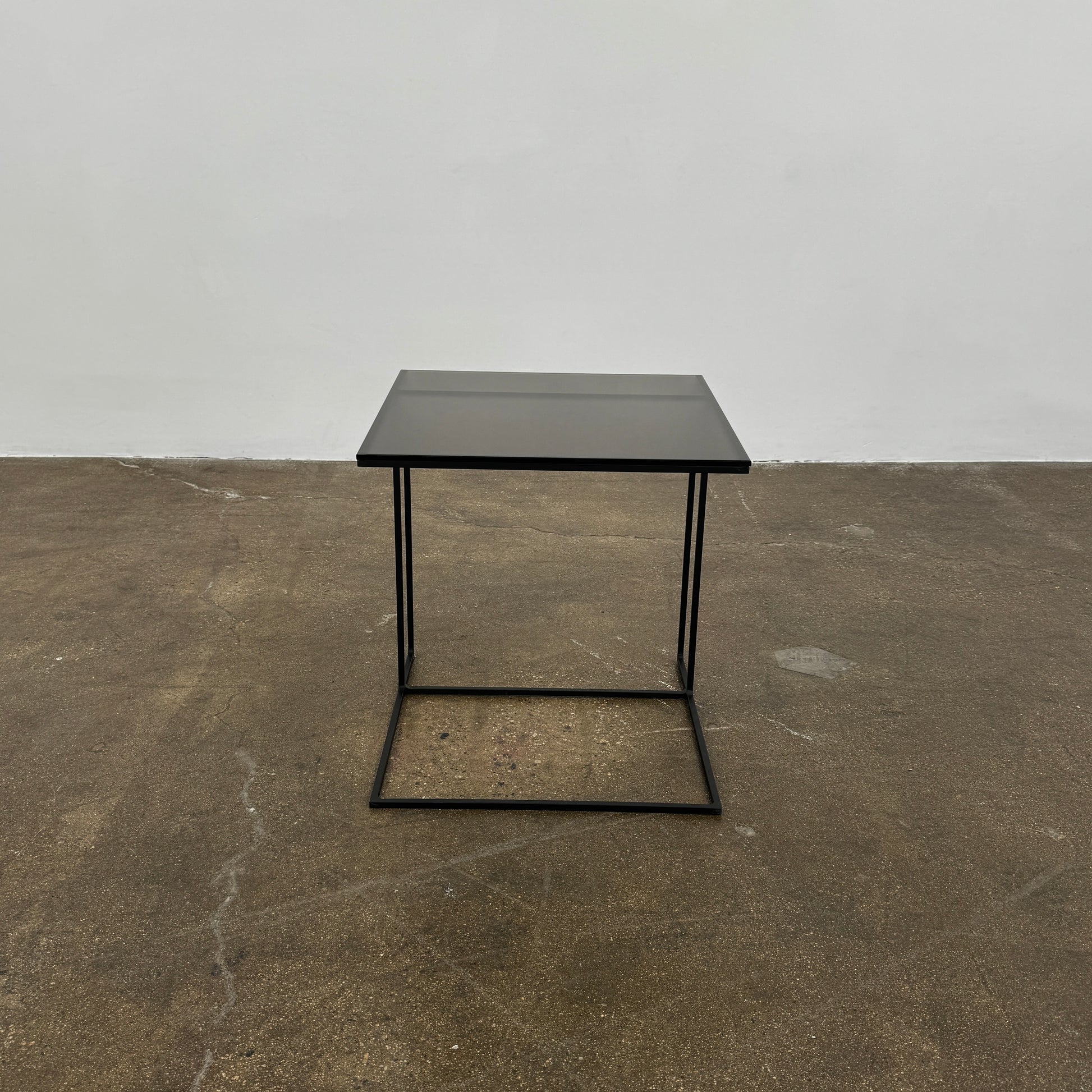 The Minotti Leger accent table by Minotti features a minimalist black metal frame with geometric lines and a flat rectangular bronze glass top, inspired by the cantilever style, shown against a plain background.
