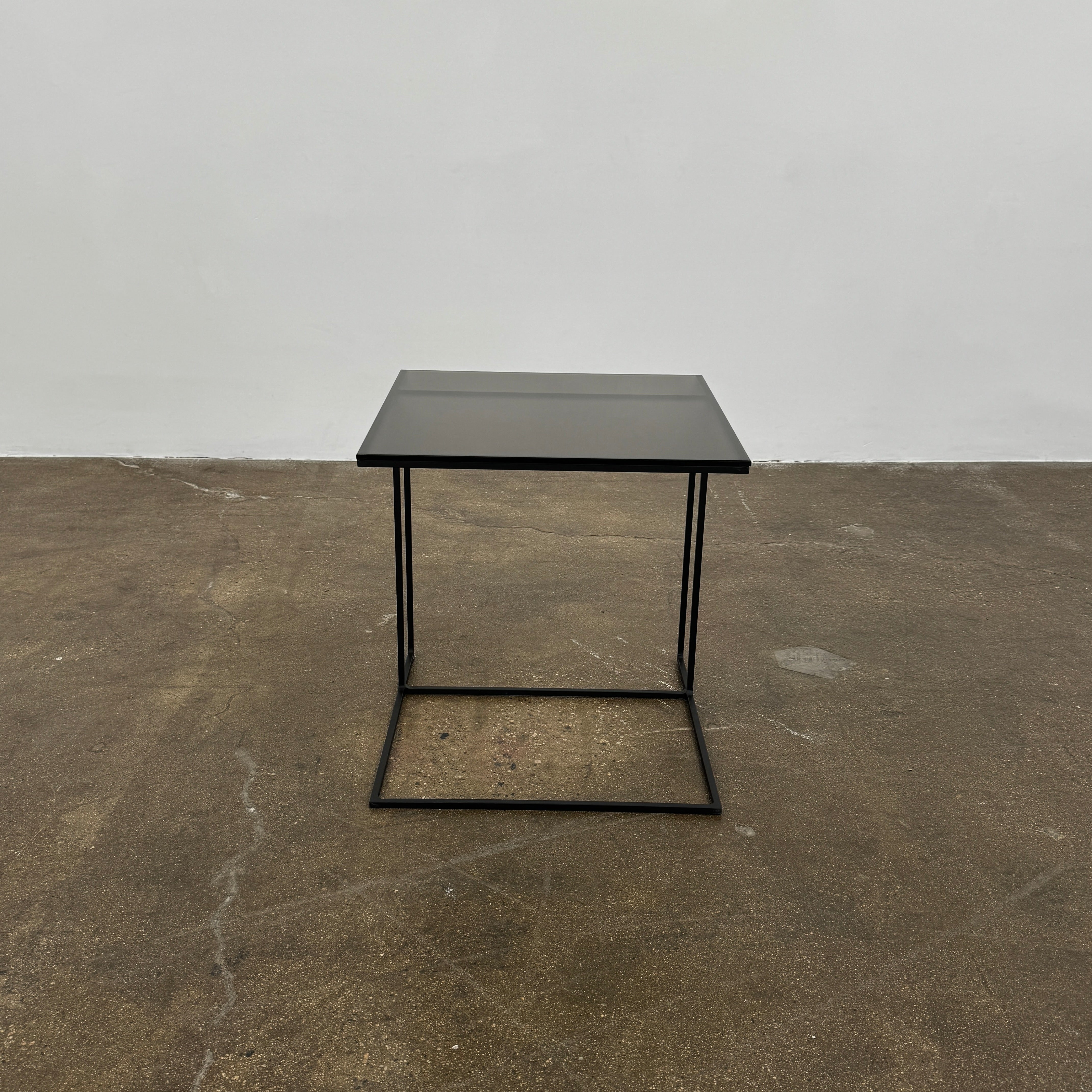 The Minotti Leger accent table by Minotti features a minimalist black metal frame with geometric lines and a flat rectangular bronze glass top, inspired by the cantilever style, shown against a plain background.