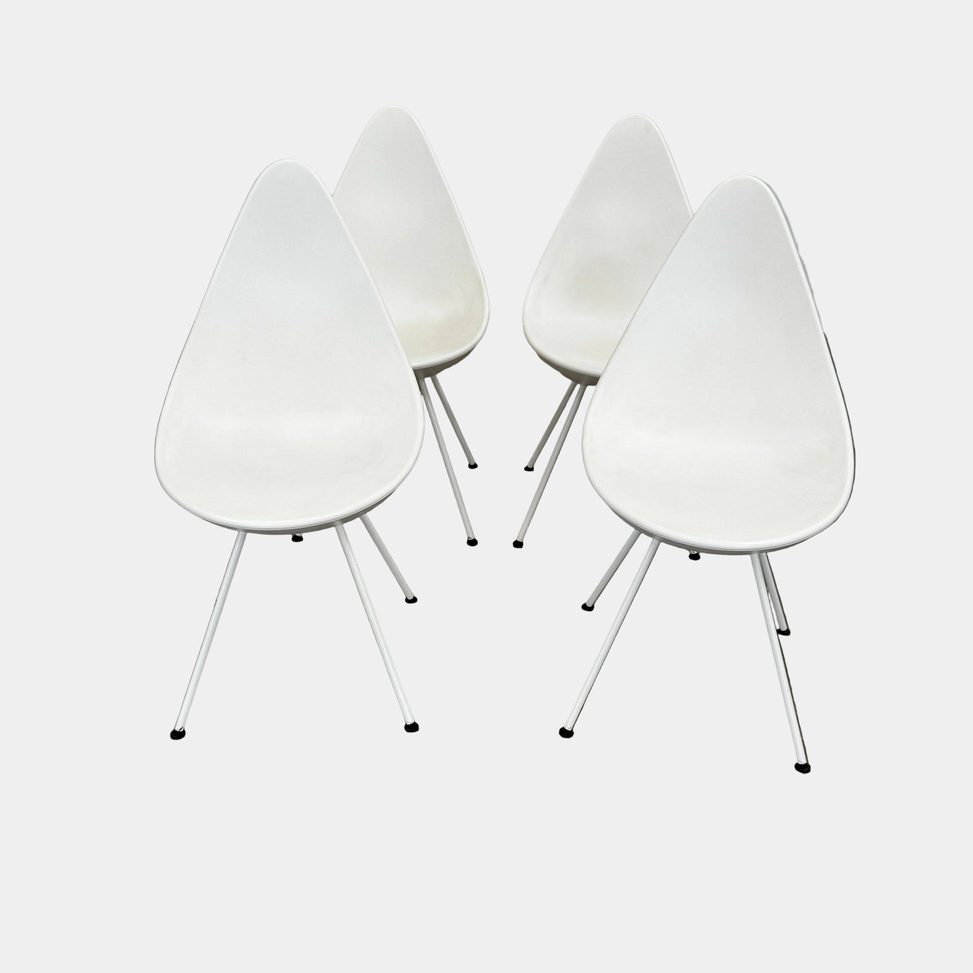 The Fritz Hansen Drop Chair Set of 4 by Fritz Hansen features white modernist chairs with teardrop-shaped backs and slender metal legs, inspired by the iconic Arne Jacobsen design, arranged in a staggered formation on a plain background.