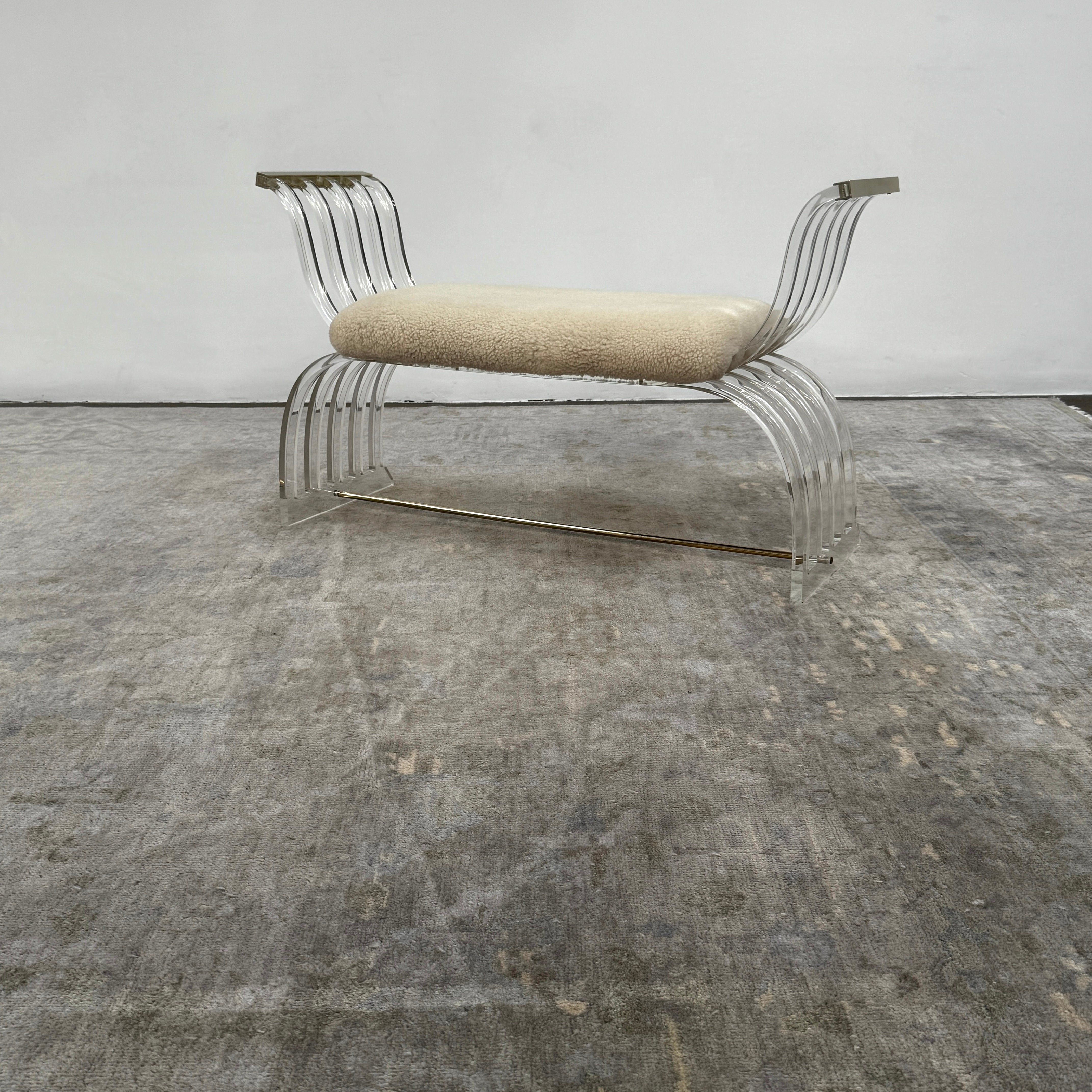 The Vintage Charles Hollis Jones Lucite Bench features a transparent acrylic frame, curved armrests, and a rectangular beige shearling cushion, shown against a plain white background.