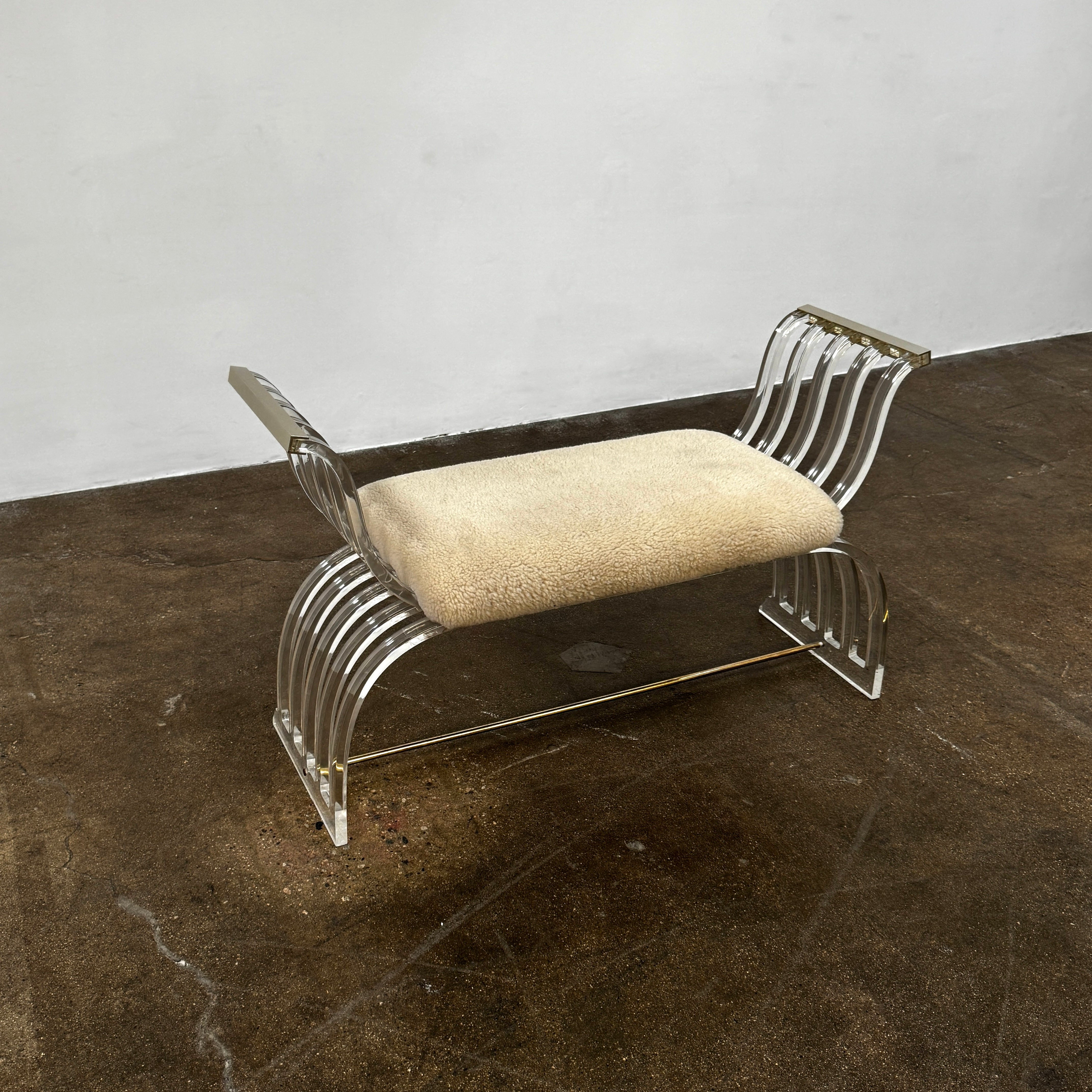 The Vintage Charles Hollis Jones Lucite Bench features a transparent acrylic frame, curved armrests, and a rectangular beige shearling cushion, shown against a plain white background.