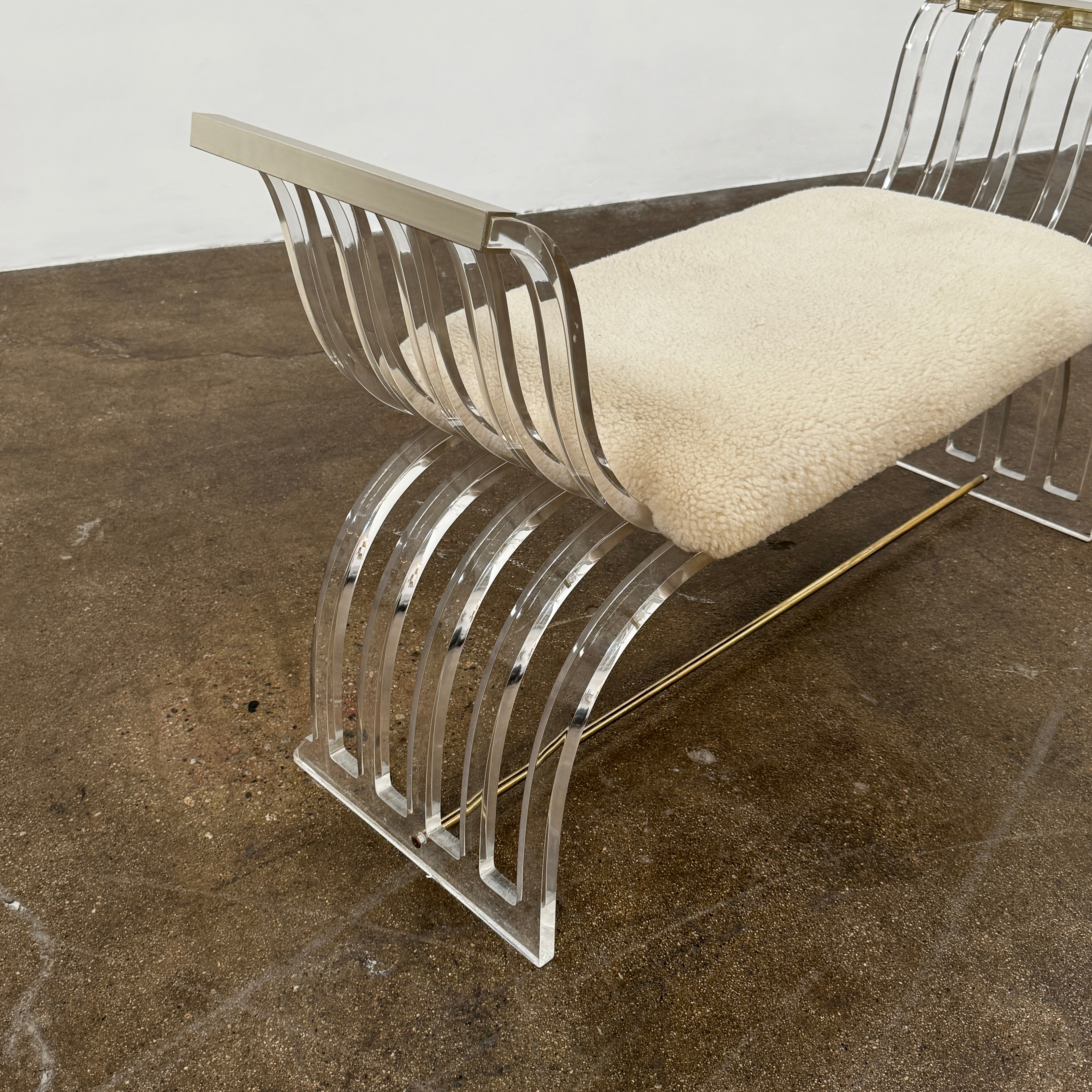 The Vintage Charles Hollis Jones Lucite Bench features a transparent acrylic frame, curved armrests, and a rectangular beige shearling cushion, shown against a plain white background.