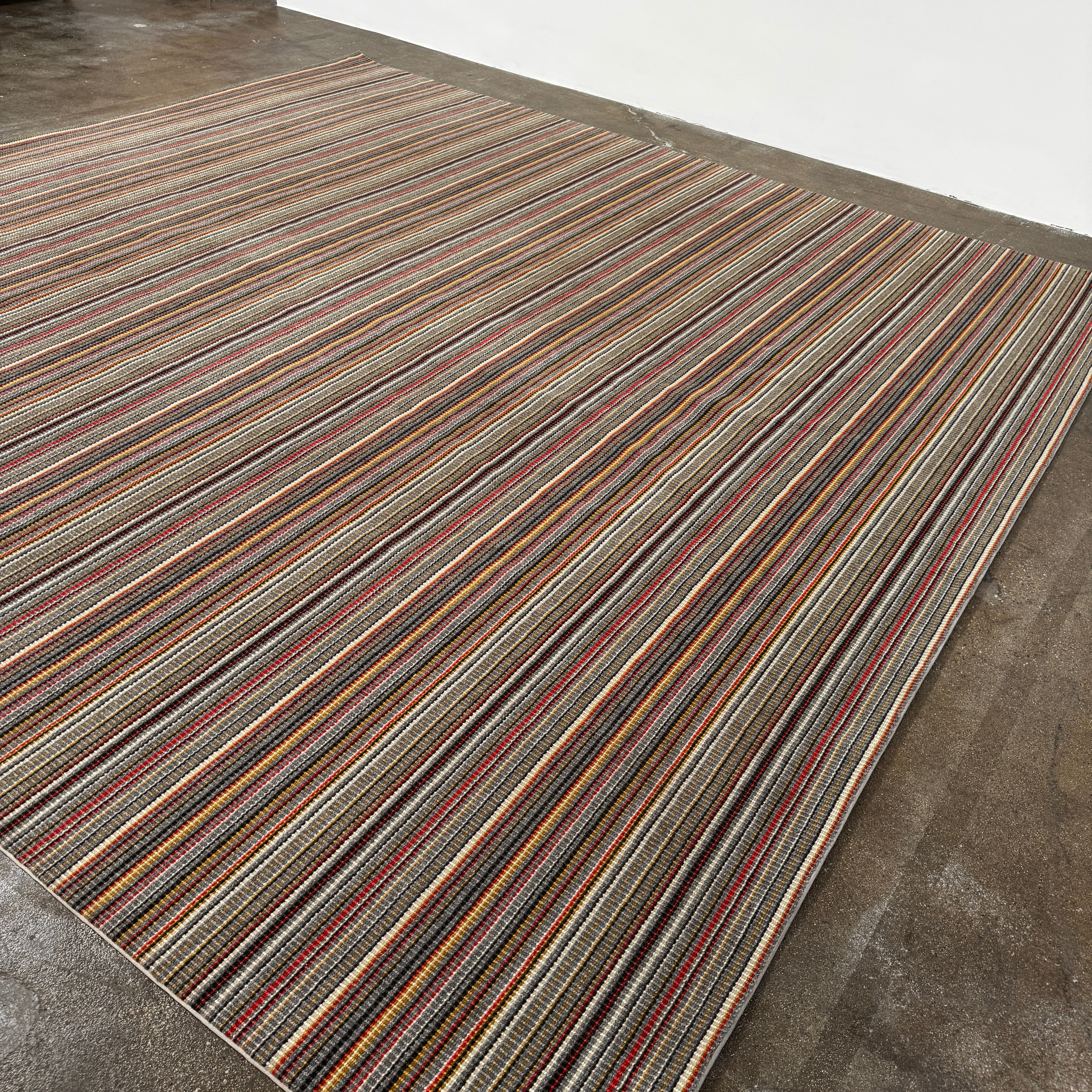 A close-up of the Maharam Interval Area Rug by Herman Miller, a modern New Zealand wool rug featuring gray, red, yellow, white, and brown stripes, shown on a white background.