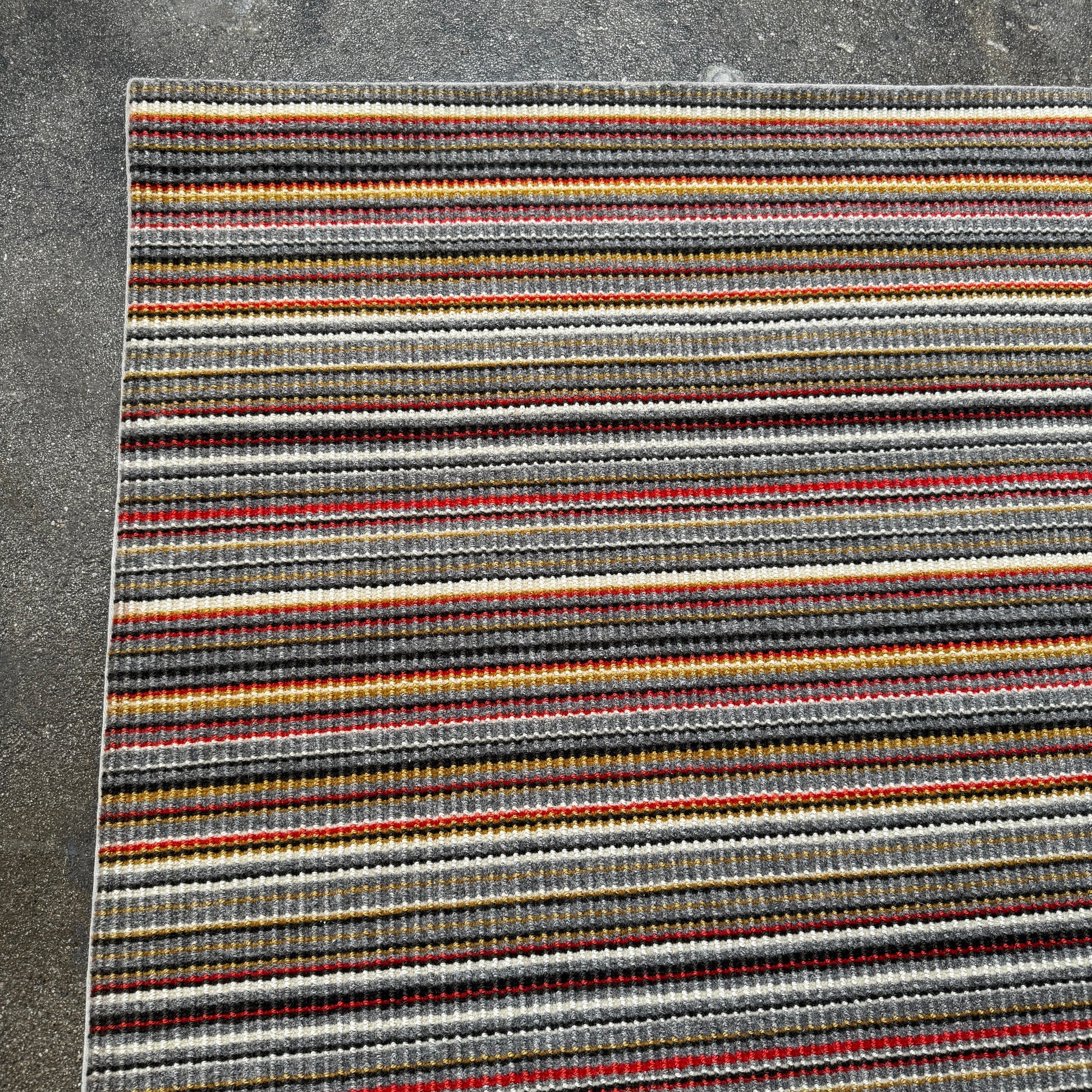 A close-up of the Maharam Interval Area Rug by Herman Miller, a modern New Zealand wool rug featuring gray, red, yellow, white, and brown stripes, shown on a white background.