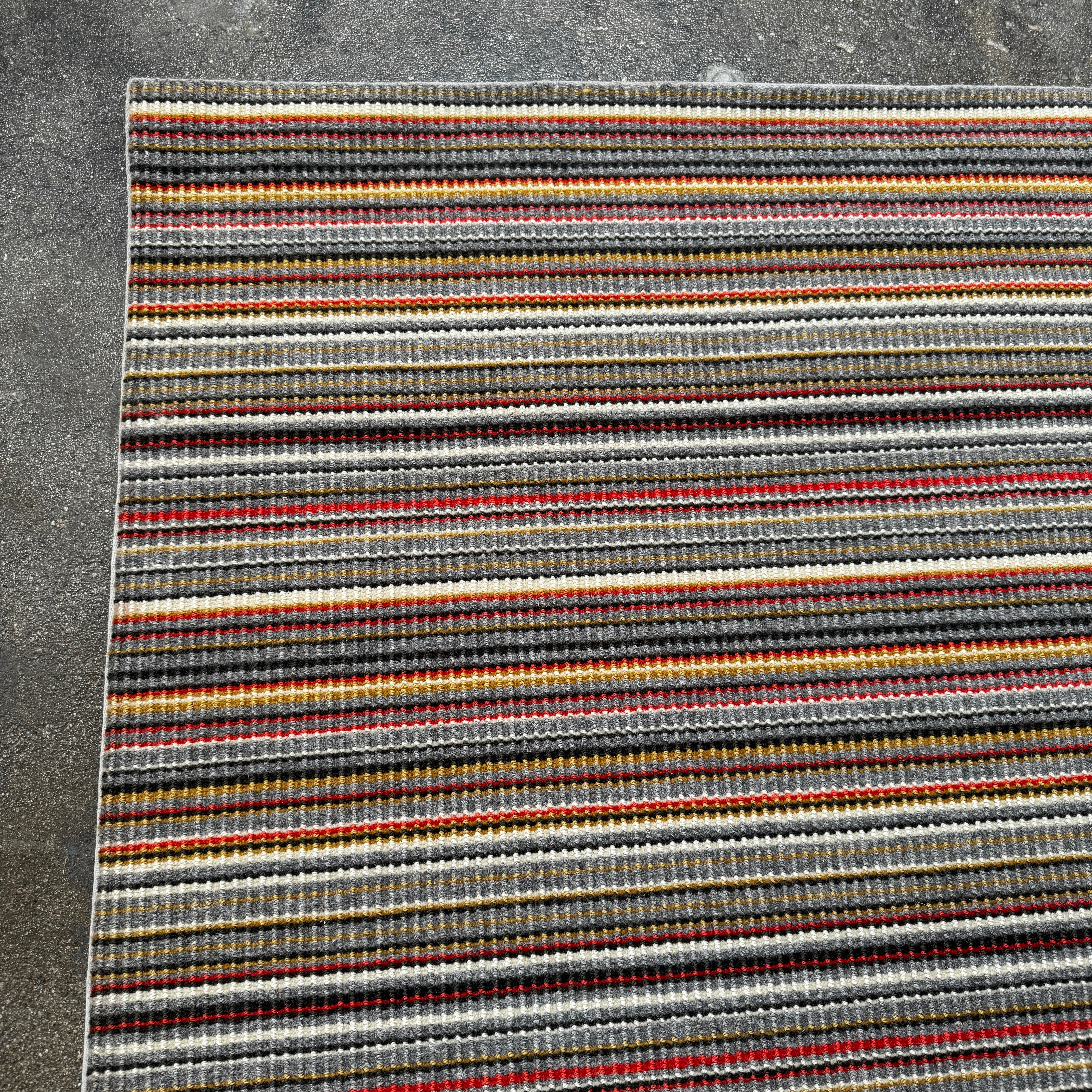 A close-up of the Maharam Interval Area Rug by Herman Miller, a modern New Zealand wool rug featuring gray, red, yellow, white, and brown stripes, shown on a white background.