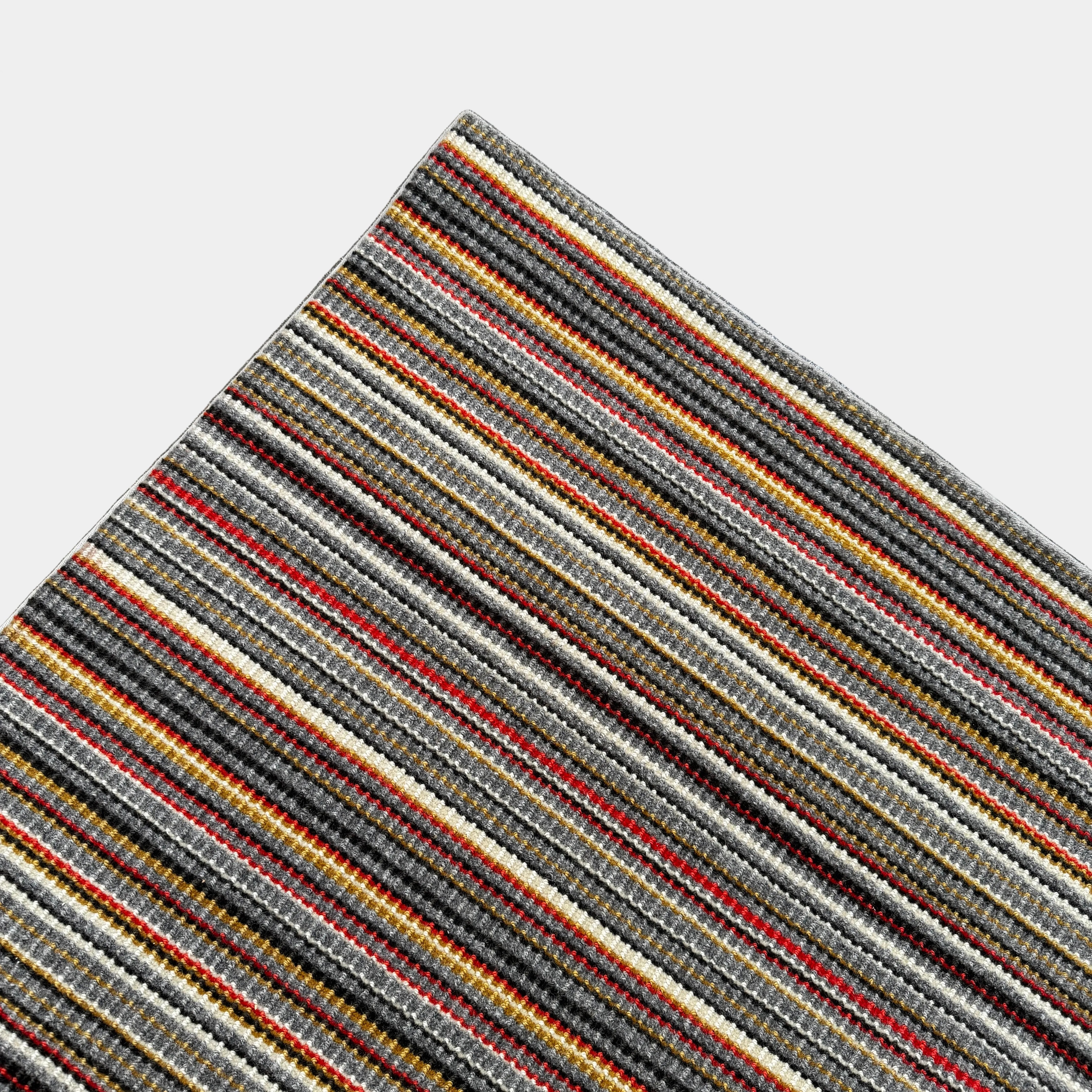A close-up of the Maharam Interval Area Rug by Herman Miller, a modern New Zealand wool rug featuring gray, red, yellow, white, and brown stripes, shown on a white background.