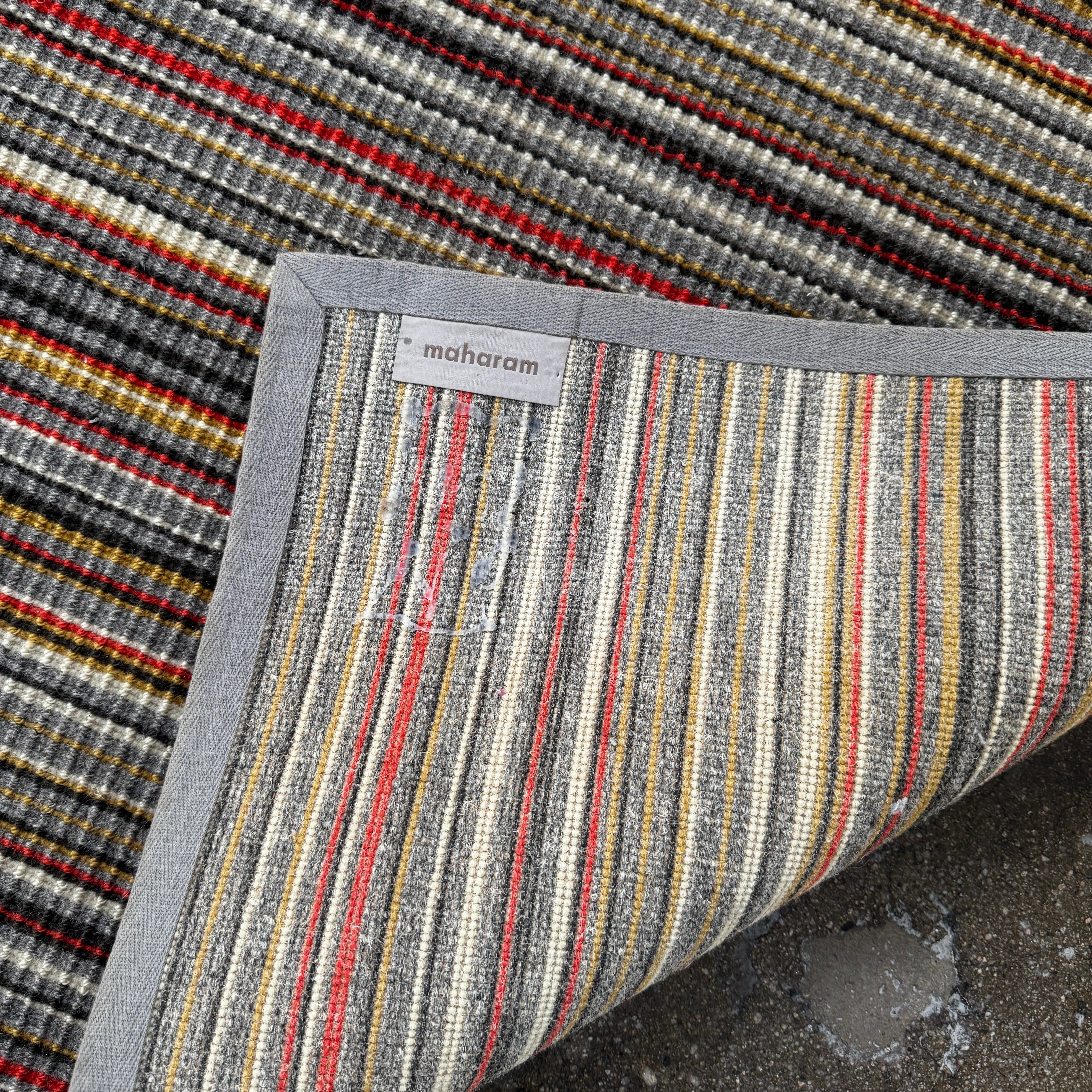 A close-up of the Maharam Interval Area Rug by Herman Miller, a modern New Zealand wool rug featuring gray, red, yellow, white, and brown stripes, shown on a white background.