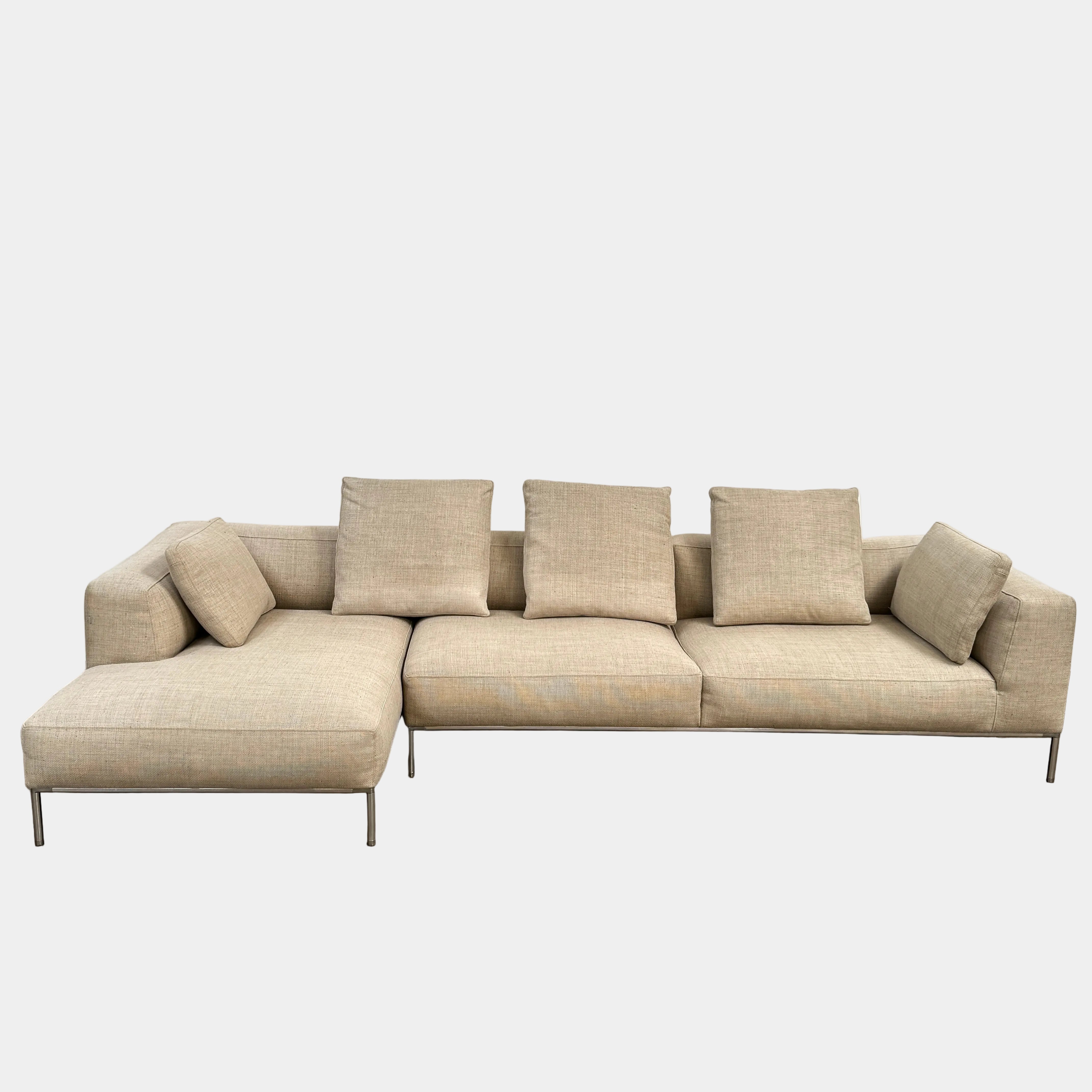 The B&B Italia Frank Sectional is a modern beige sofa featuring three large gray cushions, one beige side cushion, and sleek metal legs—an elegant statement piece from B&B Italia, set off by a plain white wall and concrete floor.