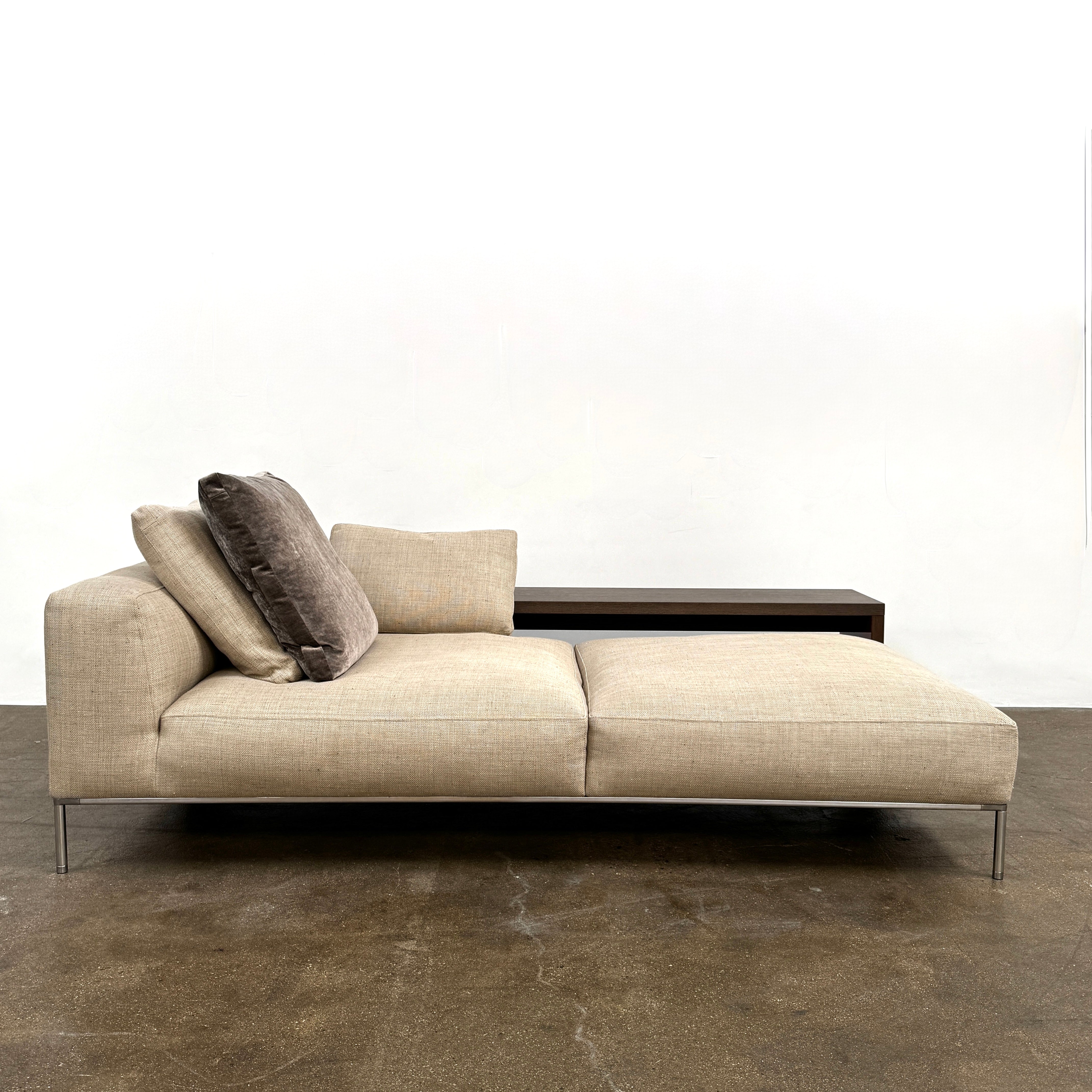 The B&B Italia Frank Chaise Lounge, upholstered in light beige with two matching cushions and metal legs, is showcased against a plain white background.
