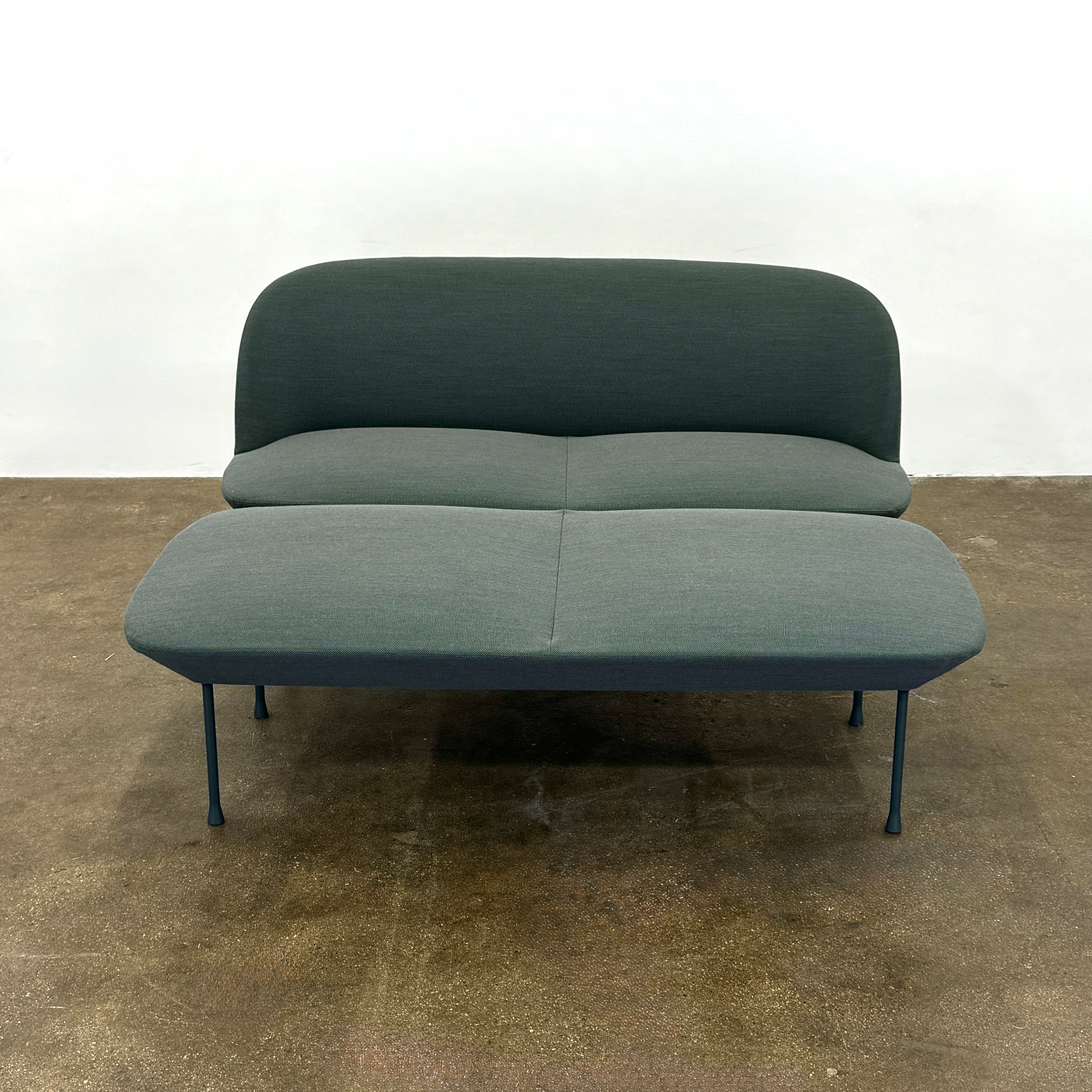 The Muuto Oslo 2-seat sofa & bench by Muuto, featuring modern green upholstery and slim black legs inspired by Danish design, stands in a bright room with beige walls and sheer white curtains.
