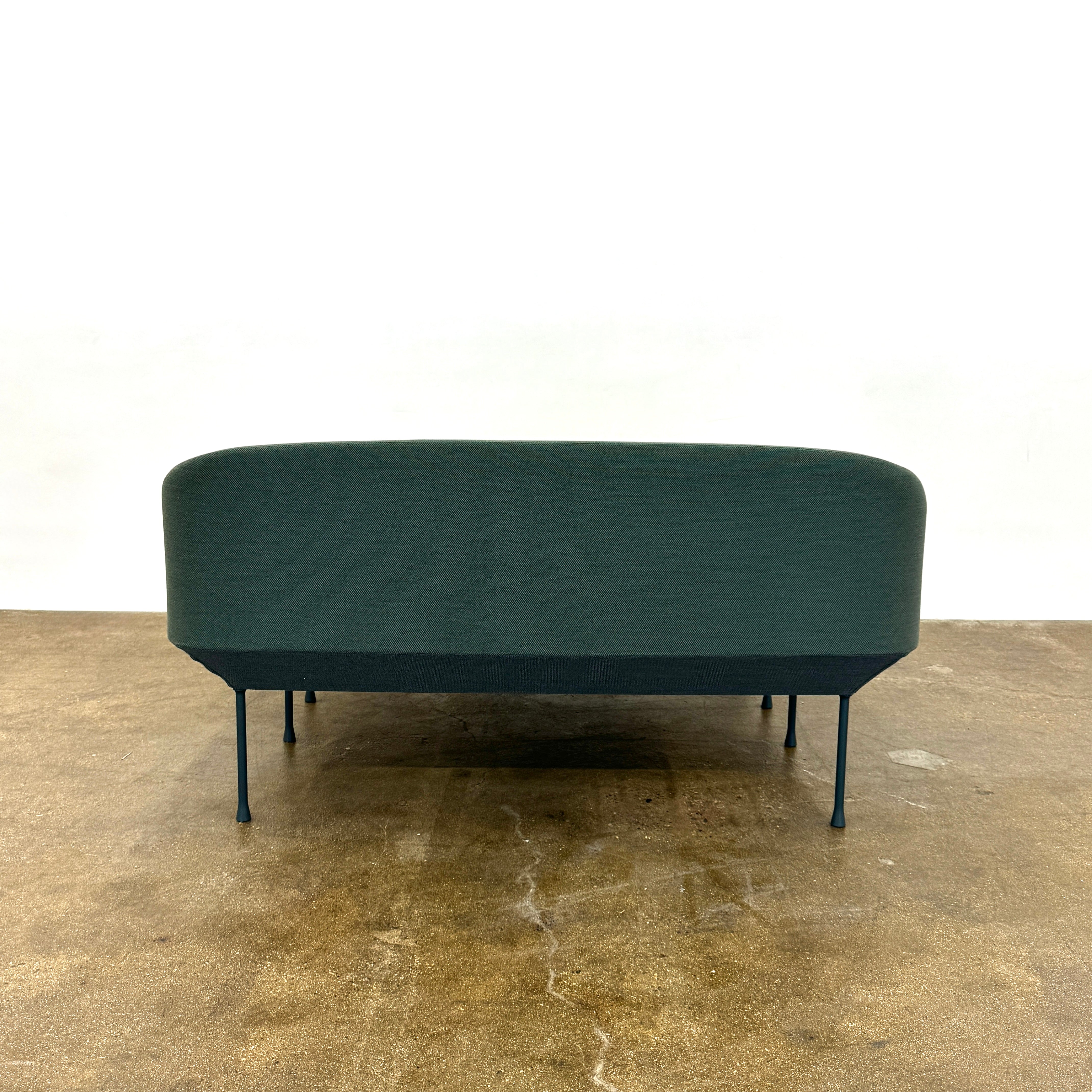 The Muuto Oslo 2-seat sofa & bench by Muuto, featuring modern green upholstery and slim black legs inspired by Danish design, stands in a bright room with beige walls and sheer white curtains.
