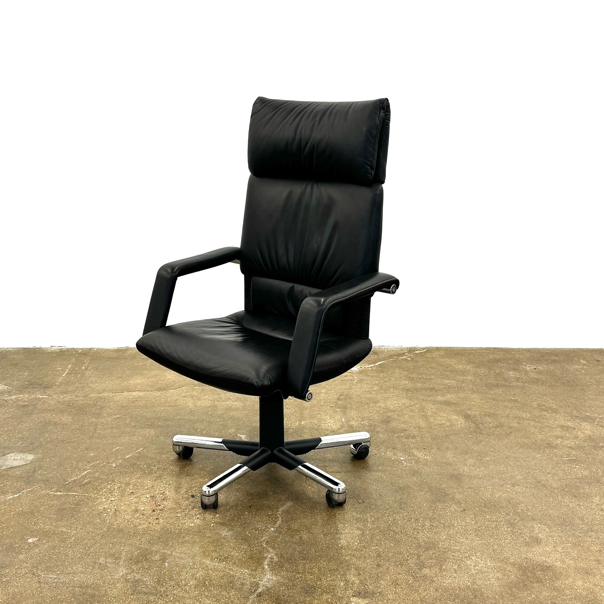 The Vitra Imago office chair by Modern Resale features black leather, armrests, a high backrest, and a metal swivel base with wheels—its sleek designer look is inspired by Mario Bellini’s timeless aesthetic and sits against a plain white background.