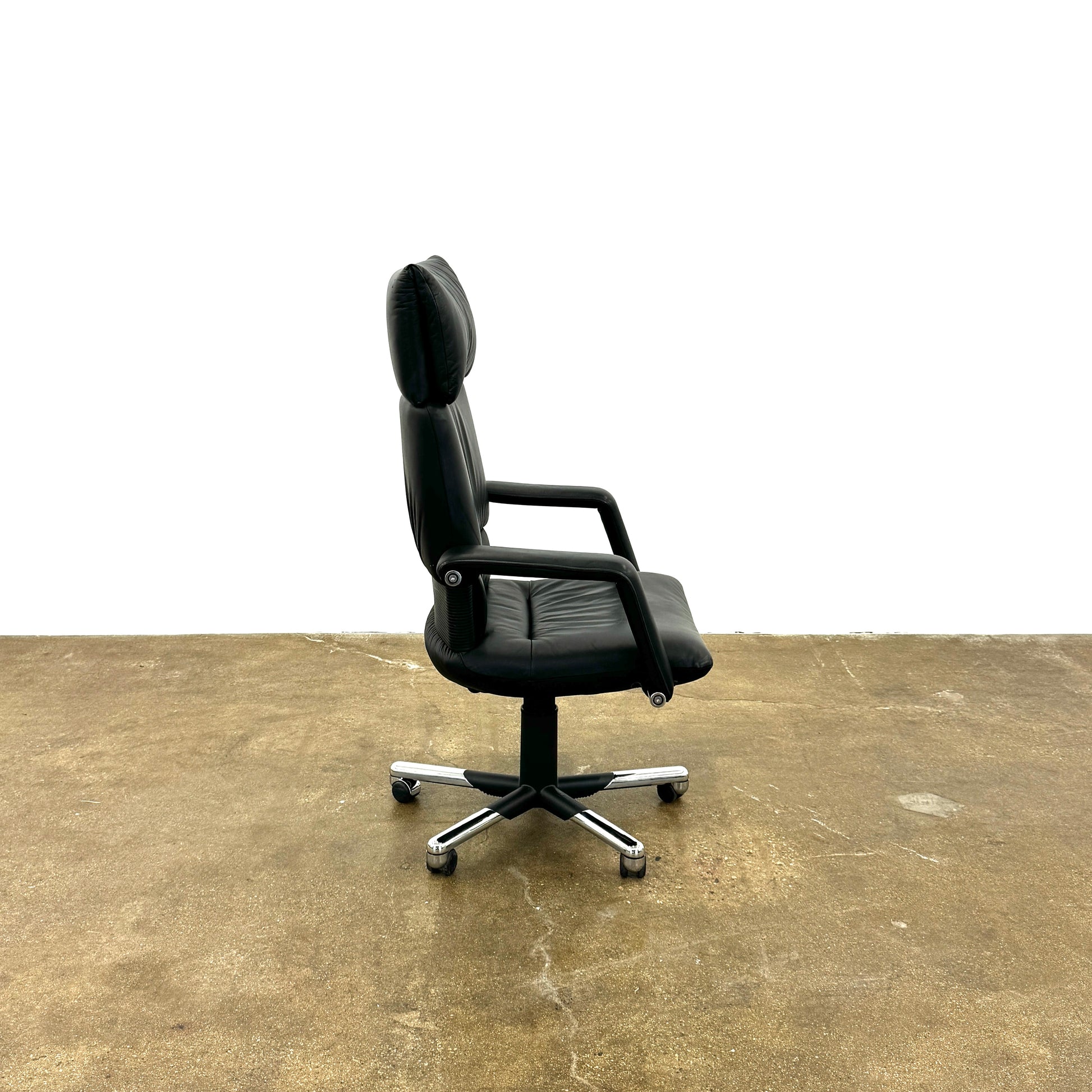 The Vitra Imago office chair by Vitra features black leather, armrests, a high backrest, and a metal five-wheel base, shown here on a concrete floor with a white background.