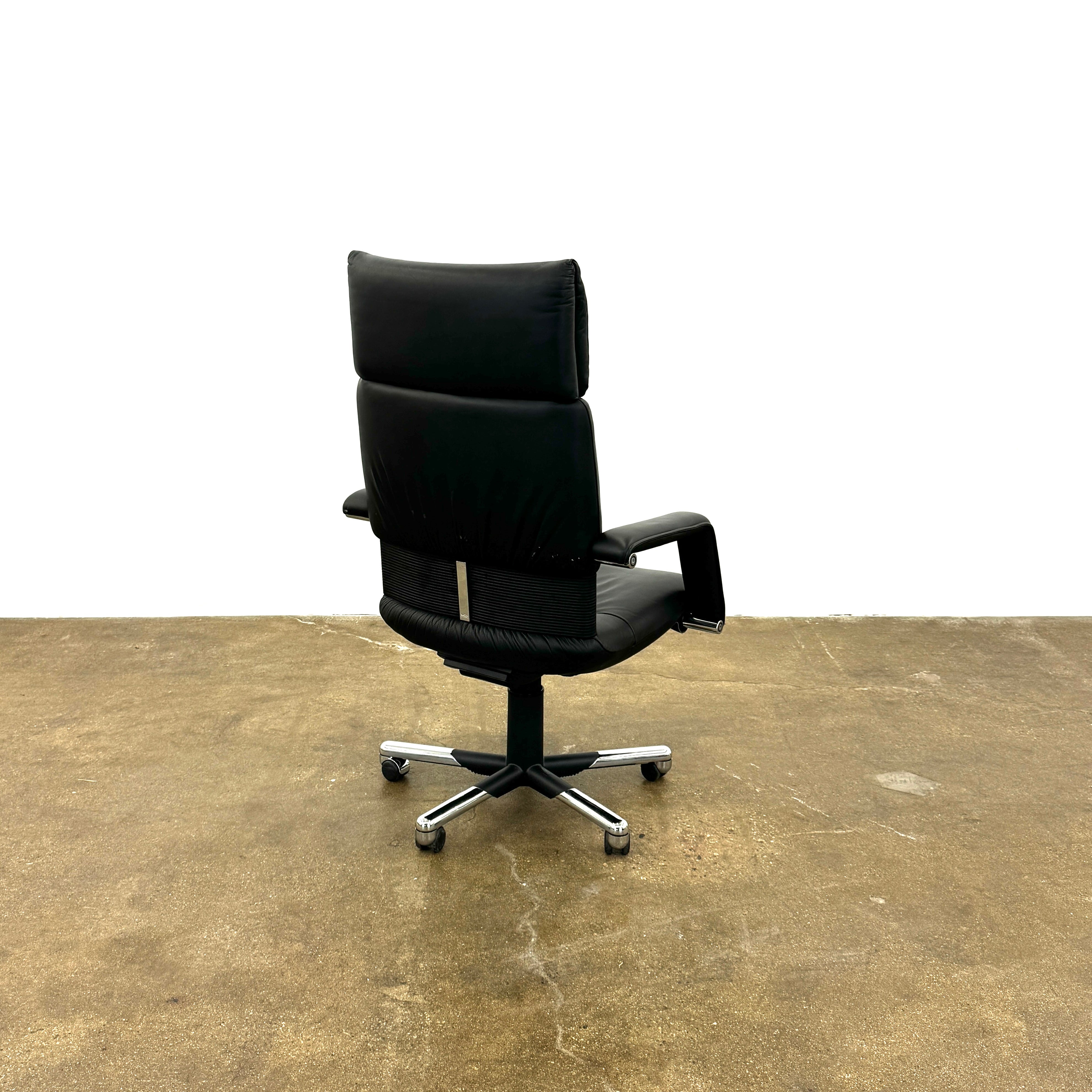 The Vitra Imago office chair by Vitra features black leather, armrests, a high backrest, and a metal five-wheel base, shown here on a concrete floor with a white background.
