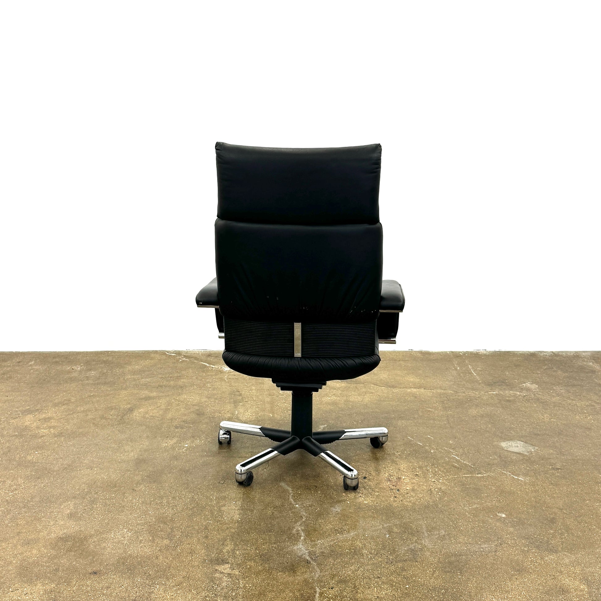The Vitra Imago office chair by Vitra features black leather, armrests, a high backrest, and a metal five-wheel base, shown here on a concrete floor with a white background.