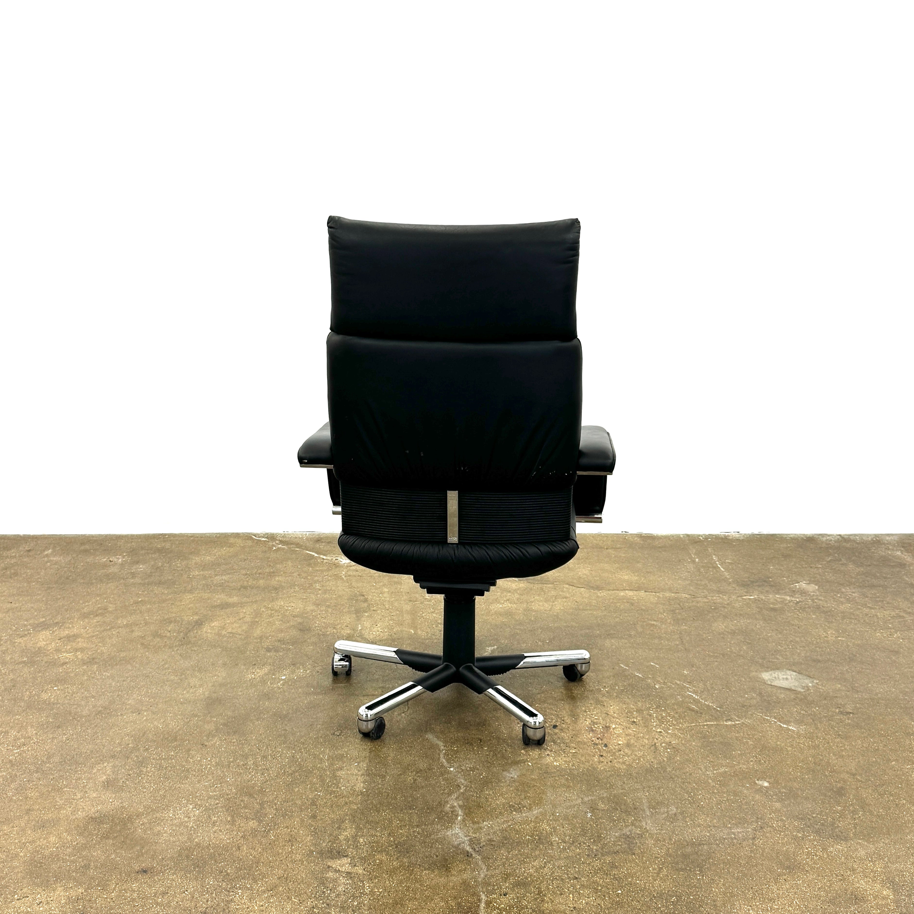 The Vitra Imago office chair by Vitra features black leather, armrests, a high backrest, and a metal five-wheel base, shown here on a concrete floor with a white background.