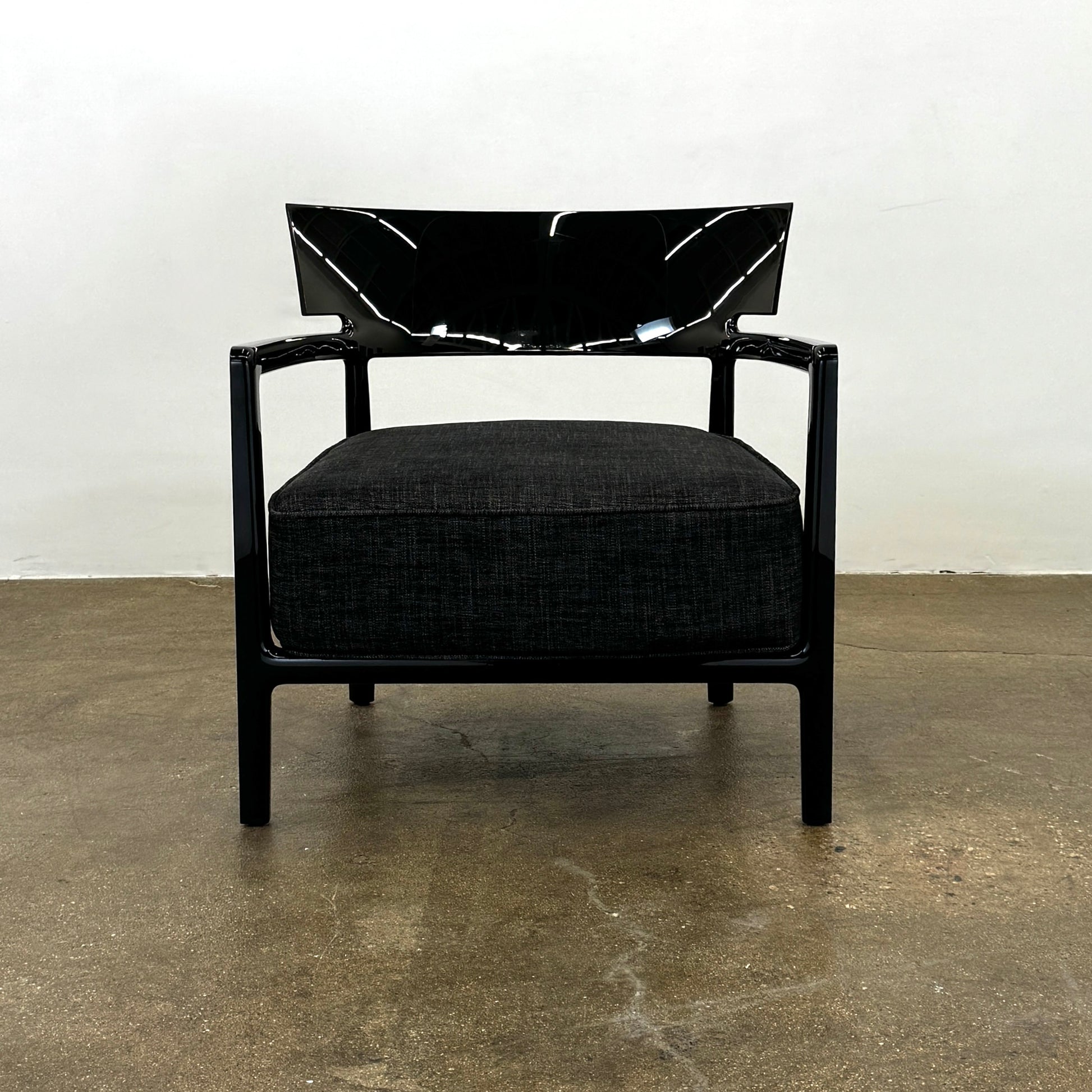 A Kartell Cara Lounge Chair by Kartell, featuring a wide black backrest and dark cushion, rests on a concrete floor in front of a plain white wall.