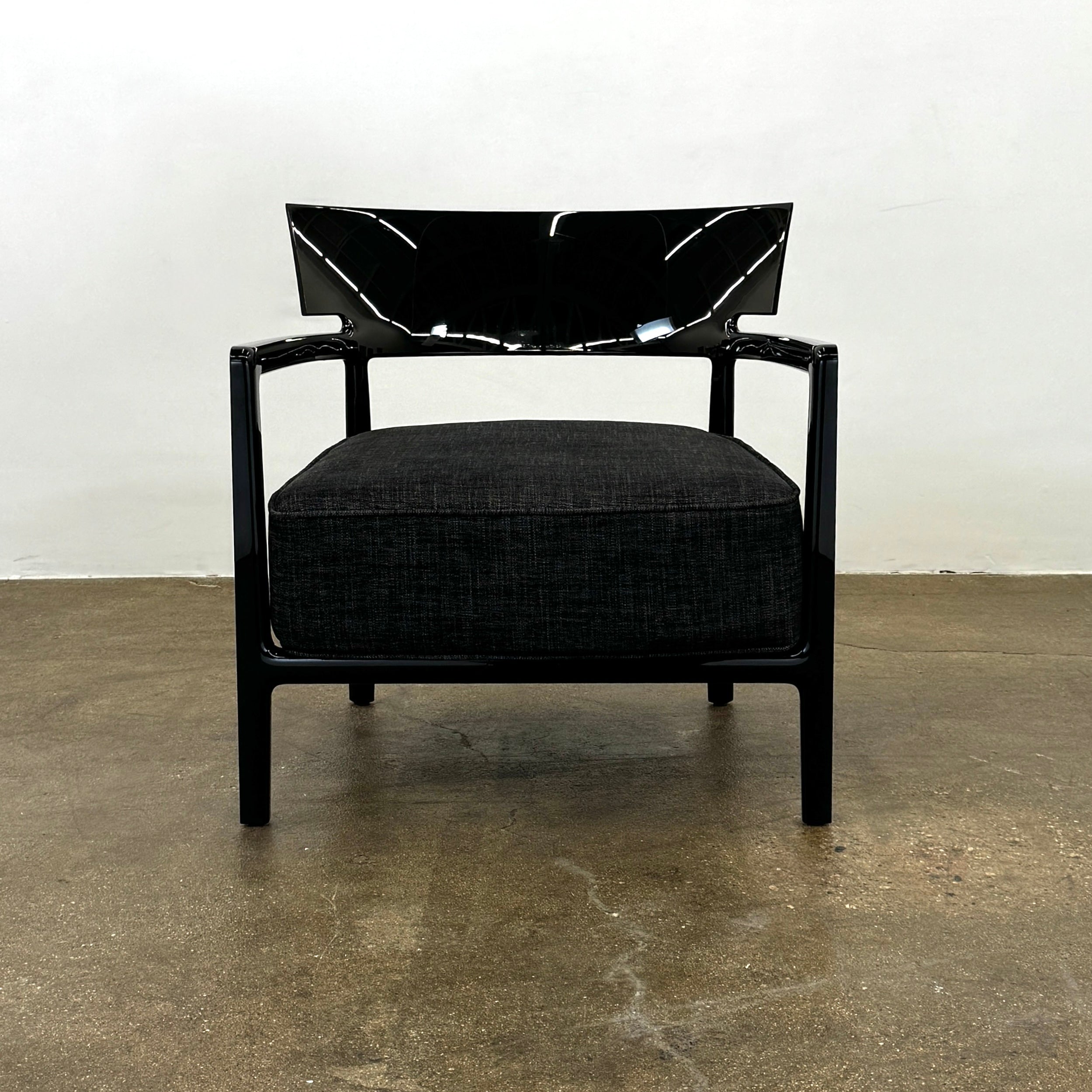 A Kartell Cara Lounge Chair by Kartell, featuring a wide black backrest and dark cushion, rests on a concrete floor in front of a plain white wall.