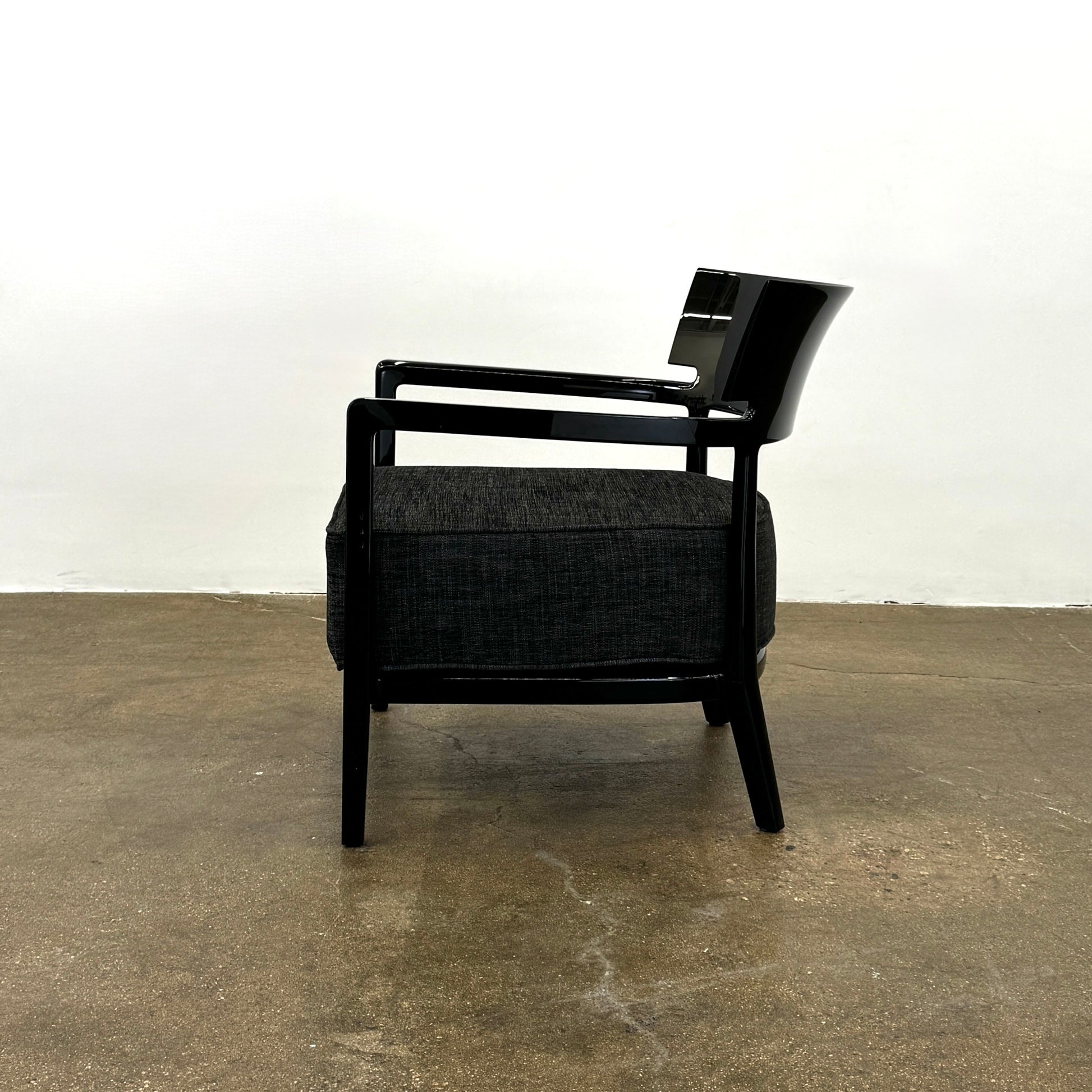 A Kartell Cara Lounge Chair by Kartell, featuring a wide black backrest and dark cushion, rests on a concrete floor in front of a plain white wall.
