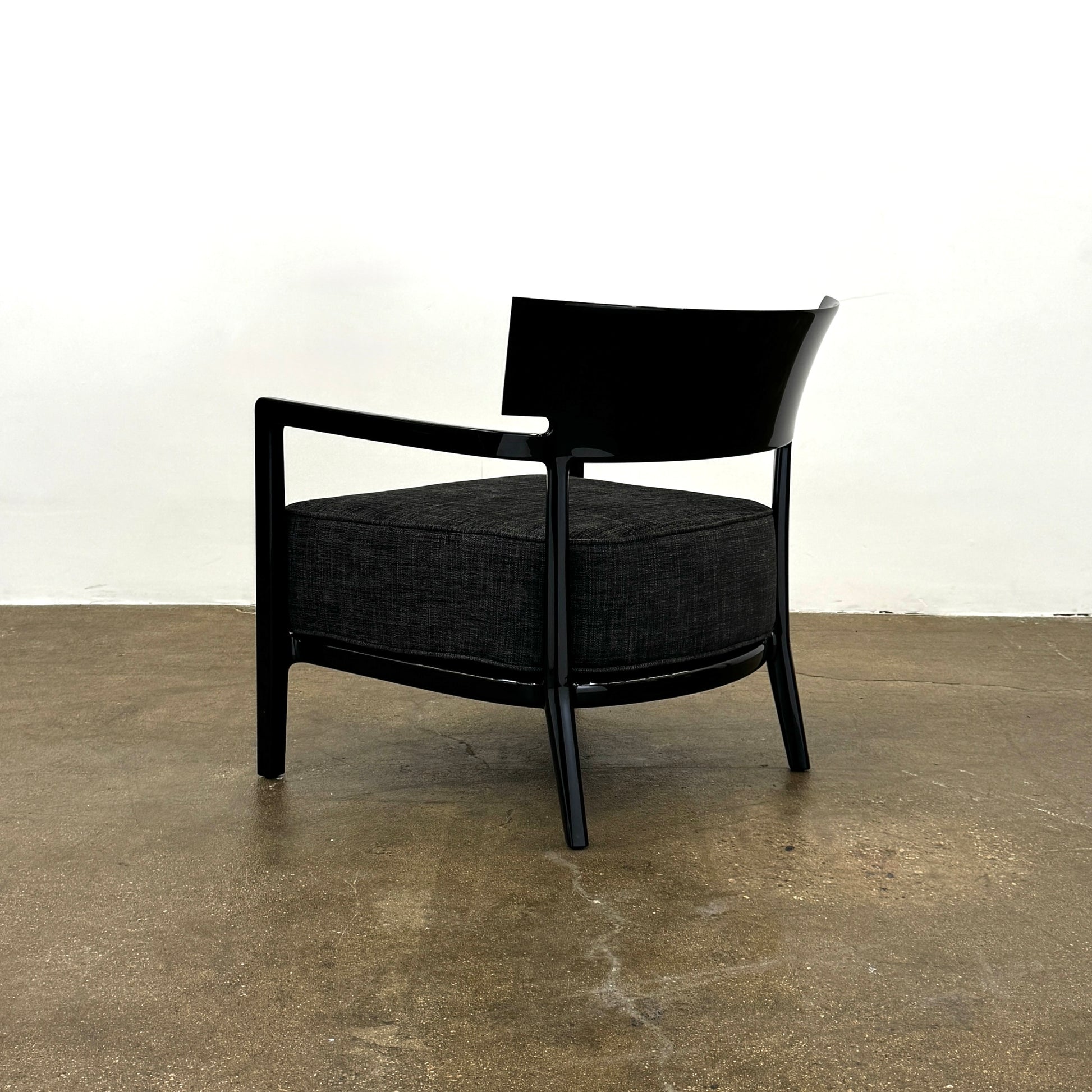 A Kartell Cara Lounge Chair by Kartell, featuring a wide black backrest and dark cushion, rests on a concrete floor in front of a plain white wall.