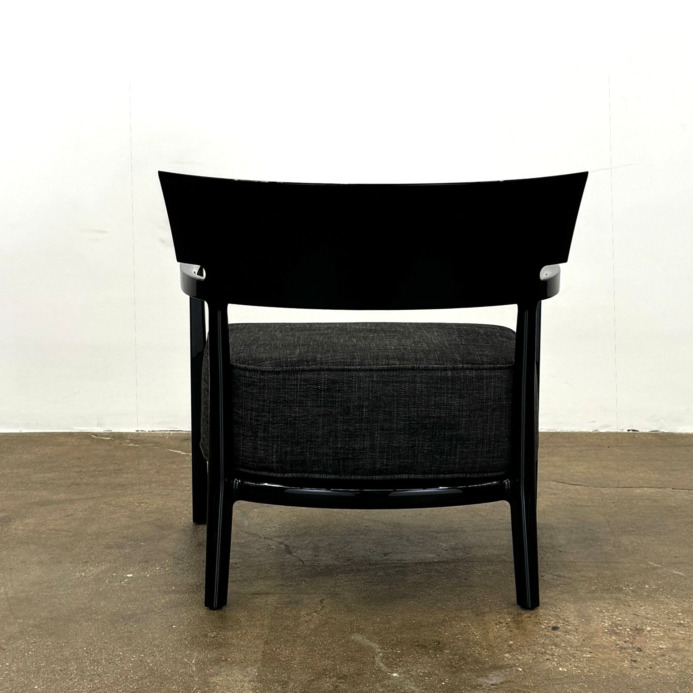 A Kartell Cara Lounge Chair by Kartell, featuring a wide black backrest and dark cushion, rests on a concrete floor in front of a plain white wall.