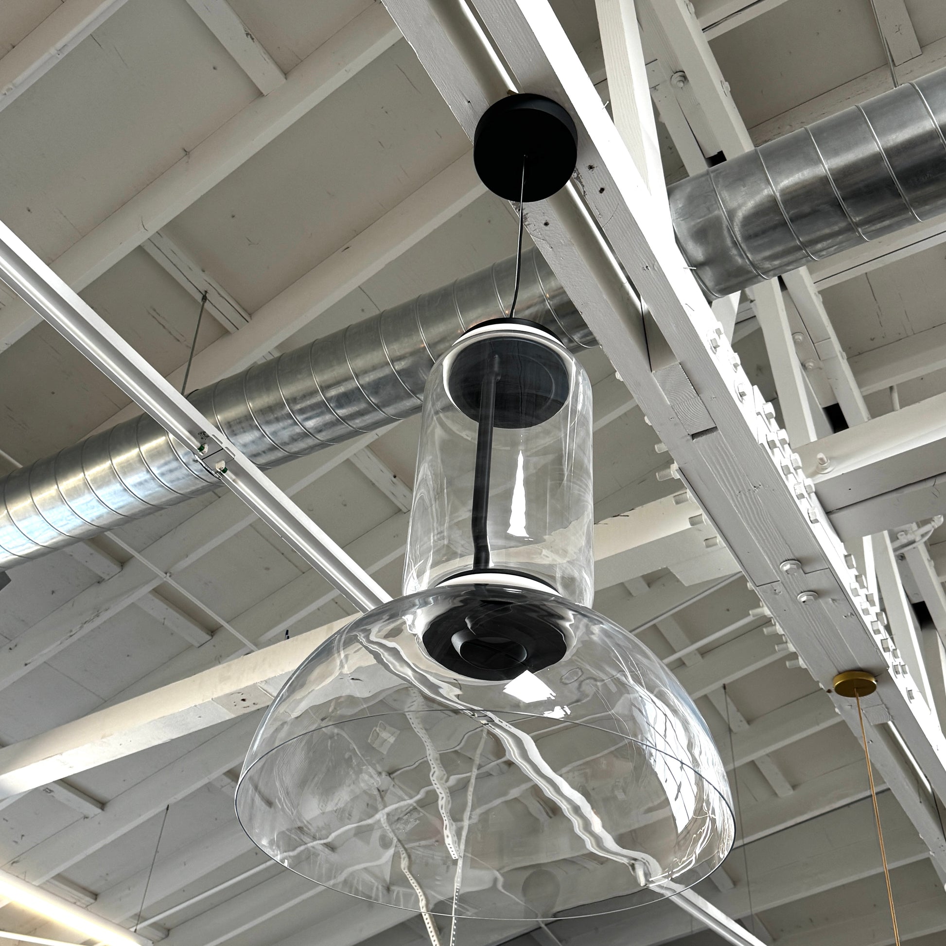The Flos Noctambule Low Cylinder and Bowl LED Pendant 1 by Flos features a transparent blown crystal glass shade with a cylindrical glass element, suspended from the ceiling by a black cord.