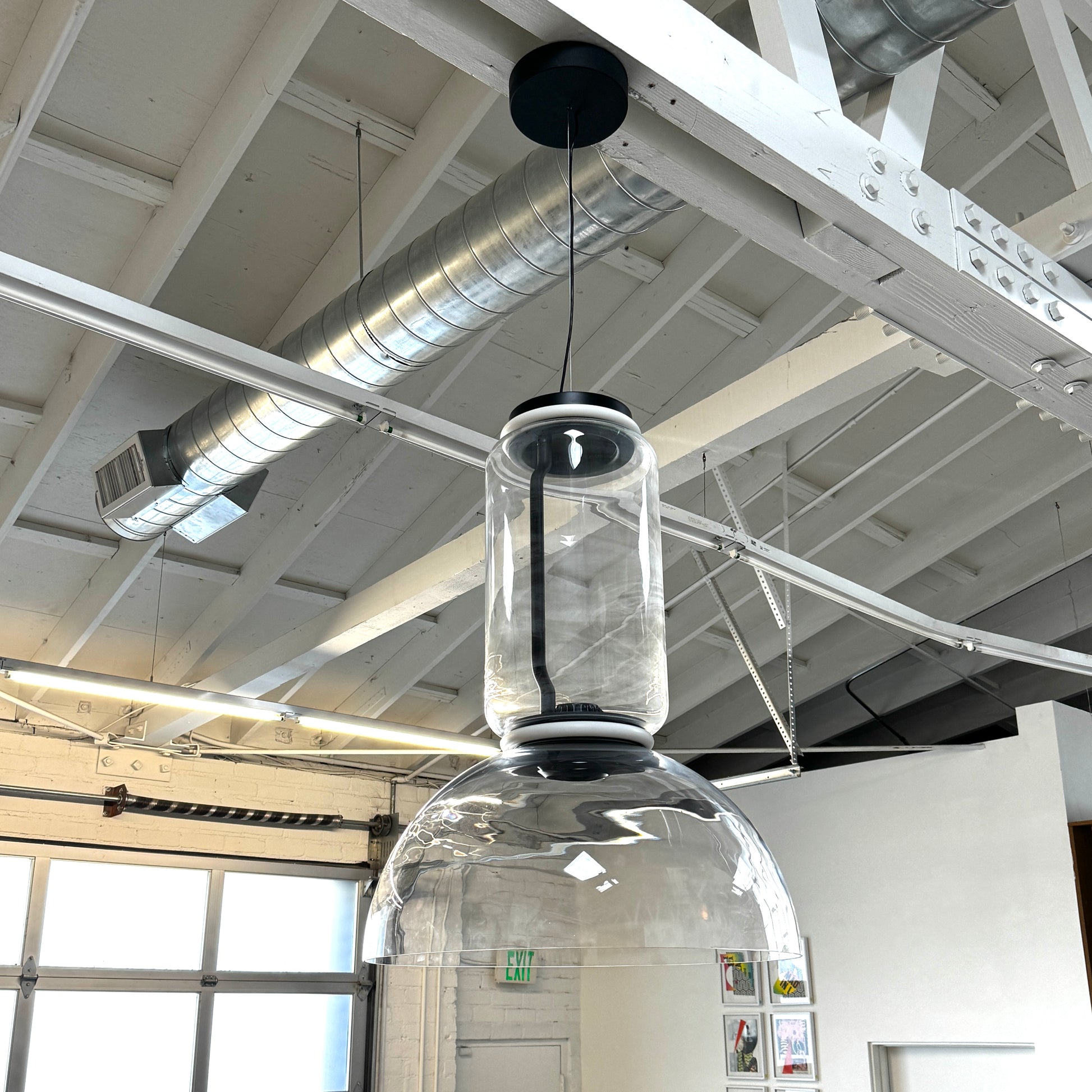 The Flos Noctambule Low Cylinder and Bowl LED Pendant 1 by Flos features a transparent blown crystal glass shade with a cylindrical glass element, suspended from the ceiling by a black cord.