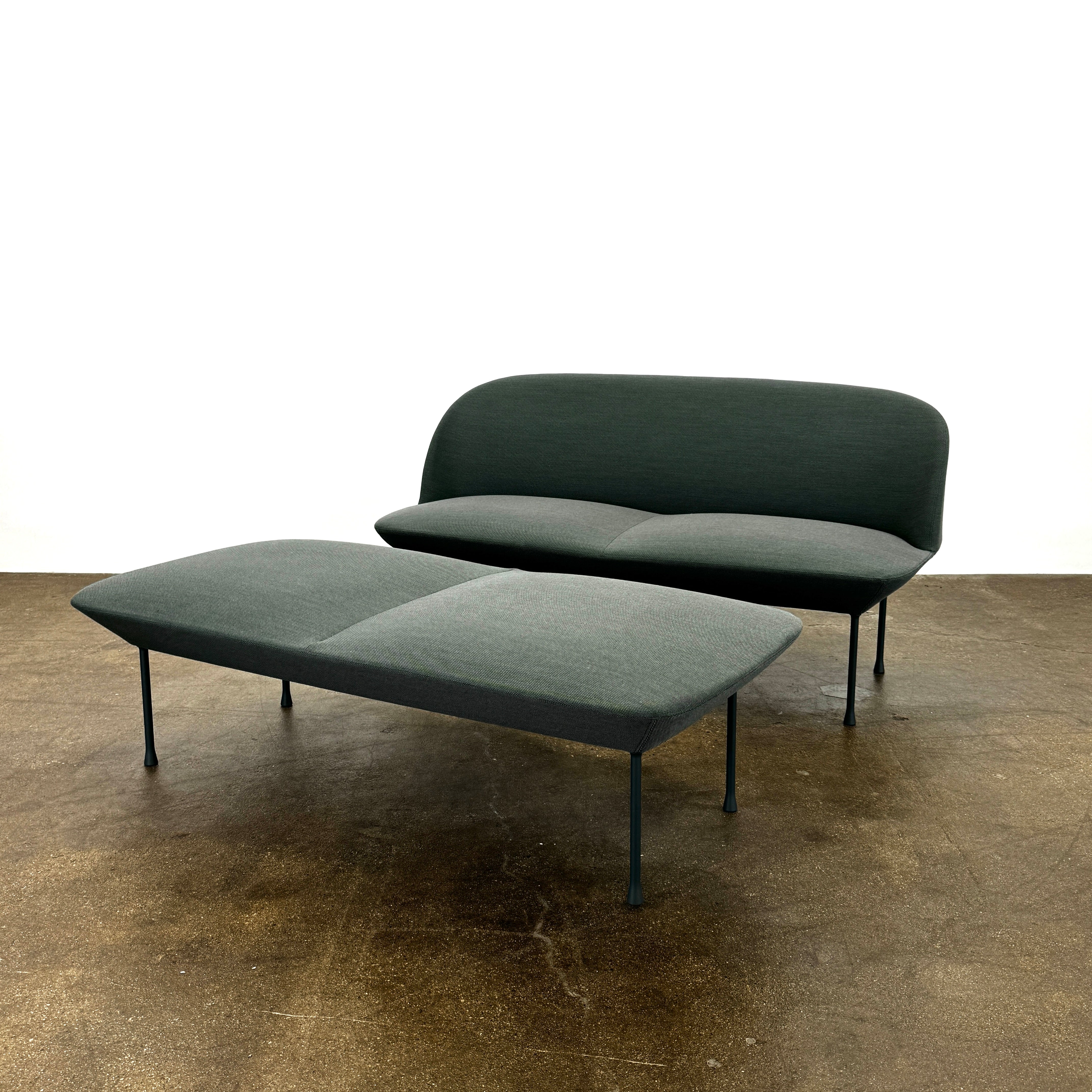 The Muuto Oslo 2-seat sofa & bench by Muuto, featuring modern green upholstery and slim black legs inspired by Danish design, stands in a bright room with beige walls and sheer white curtains.