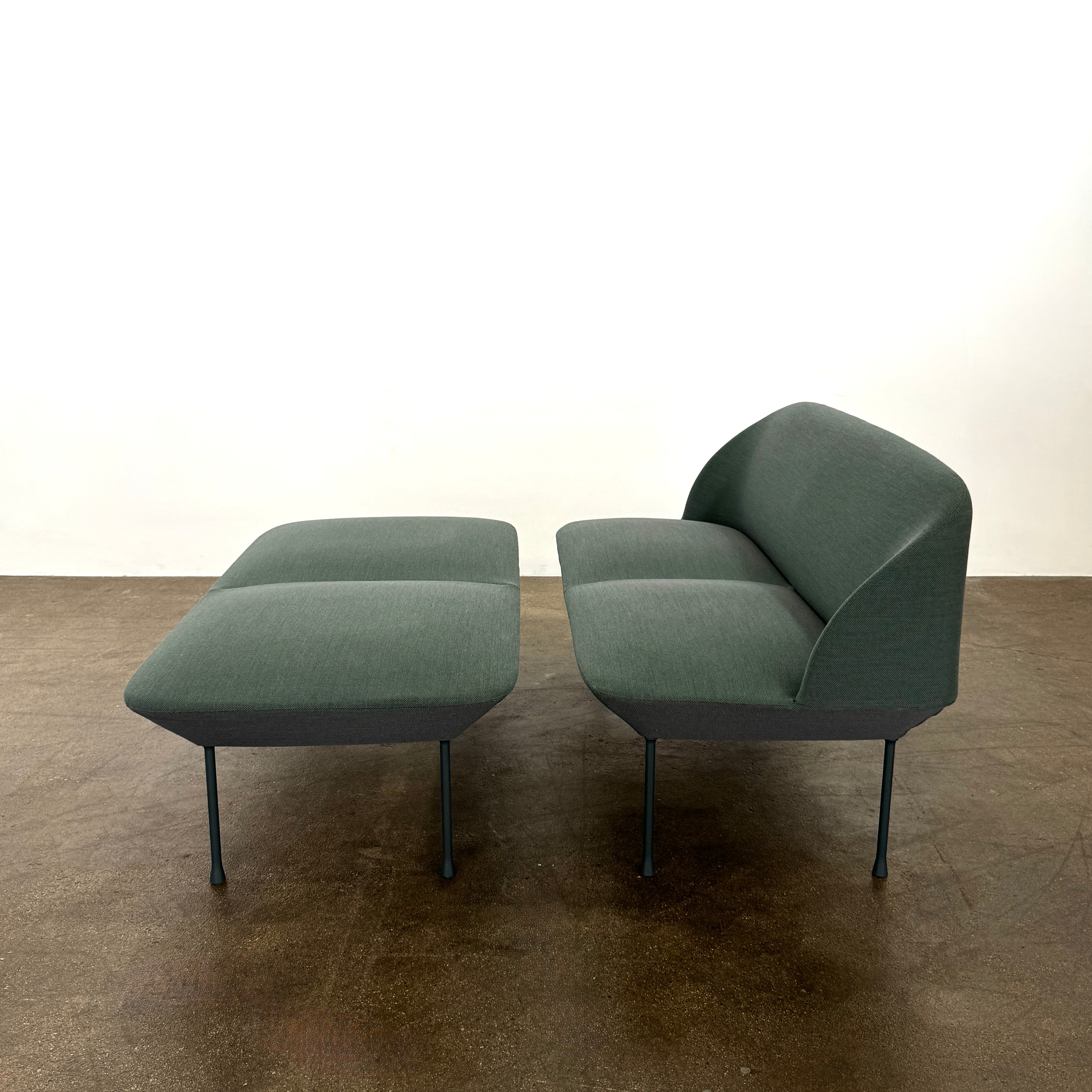 The Muuto Oslo 2-seat sofa & bench by Muuto, featuring modern green upholstery and slim black legs inspired by Danish design, stands in a bright room with beige walls and sheer white curtains.