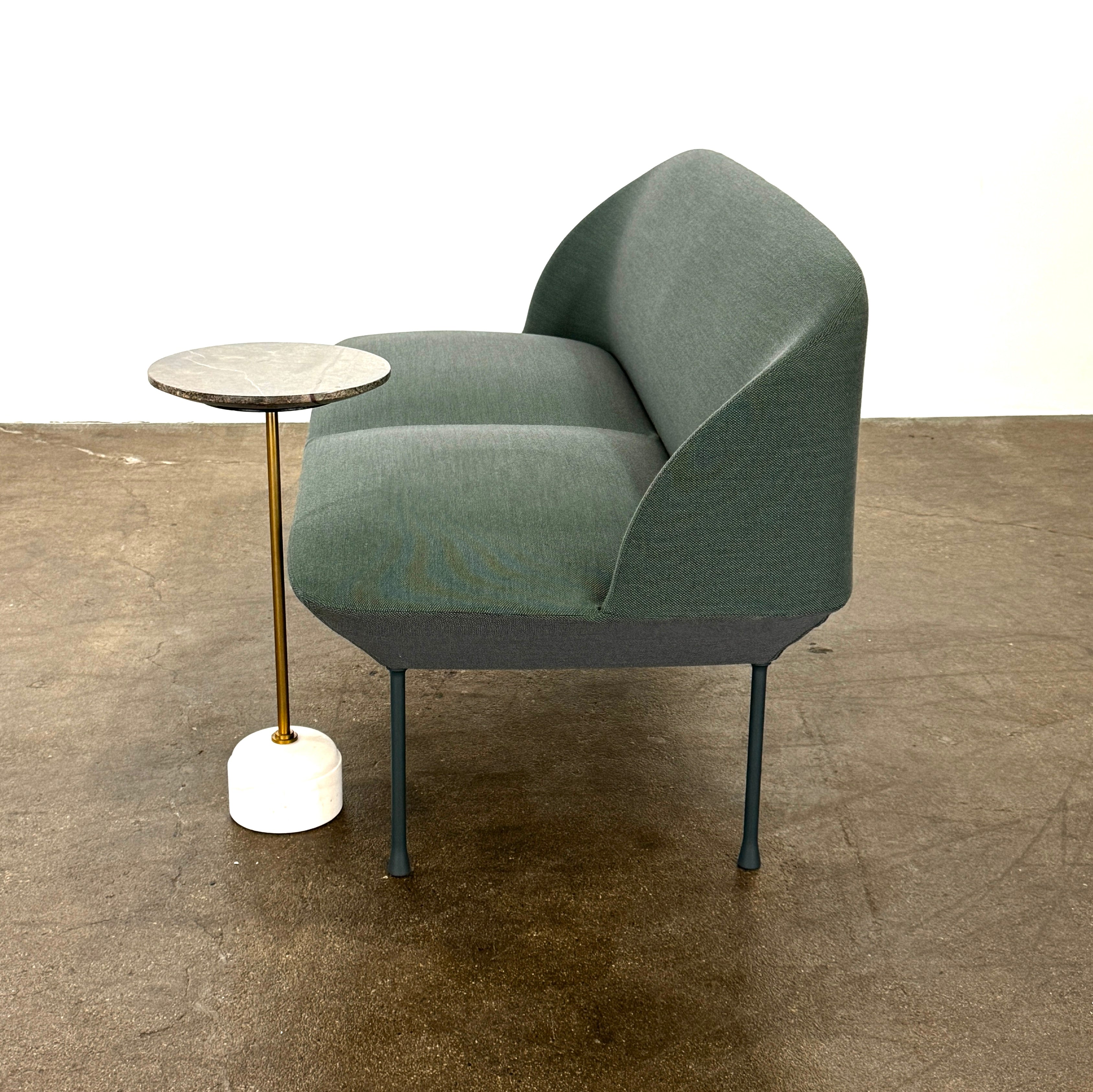 The Muuto Oslo 2-seat sofa & bench by Muuto, featuring modern green upholstery and slim black legs inspired by Danish design, stands in a bright room with beige walls and sheer white curtains.
